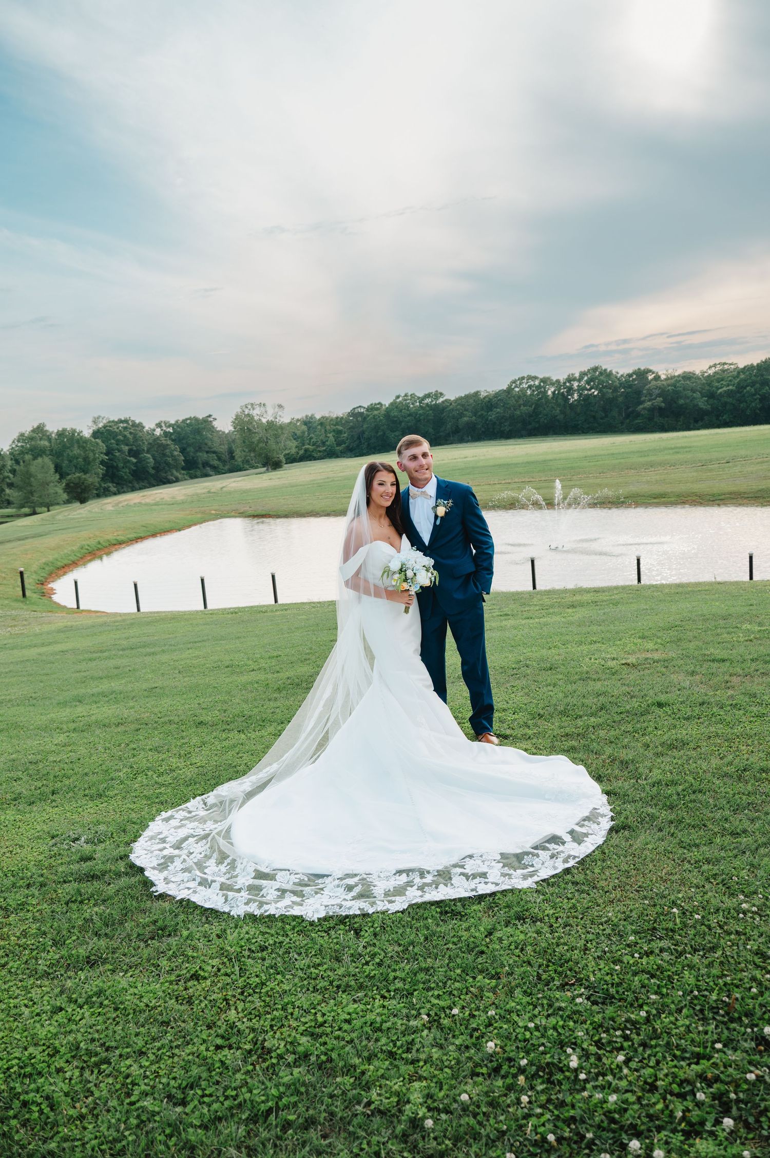 Bride in elegant white gown with lace train and groom in navy suit pose by a serene pond at golden hour.