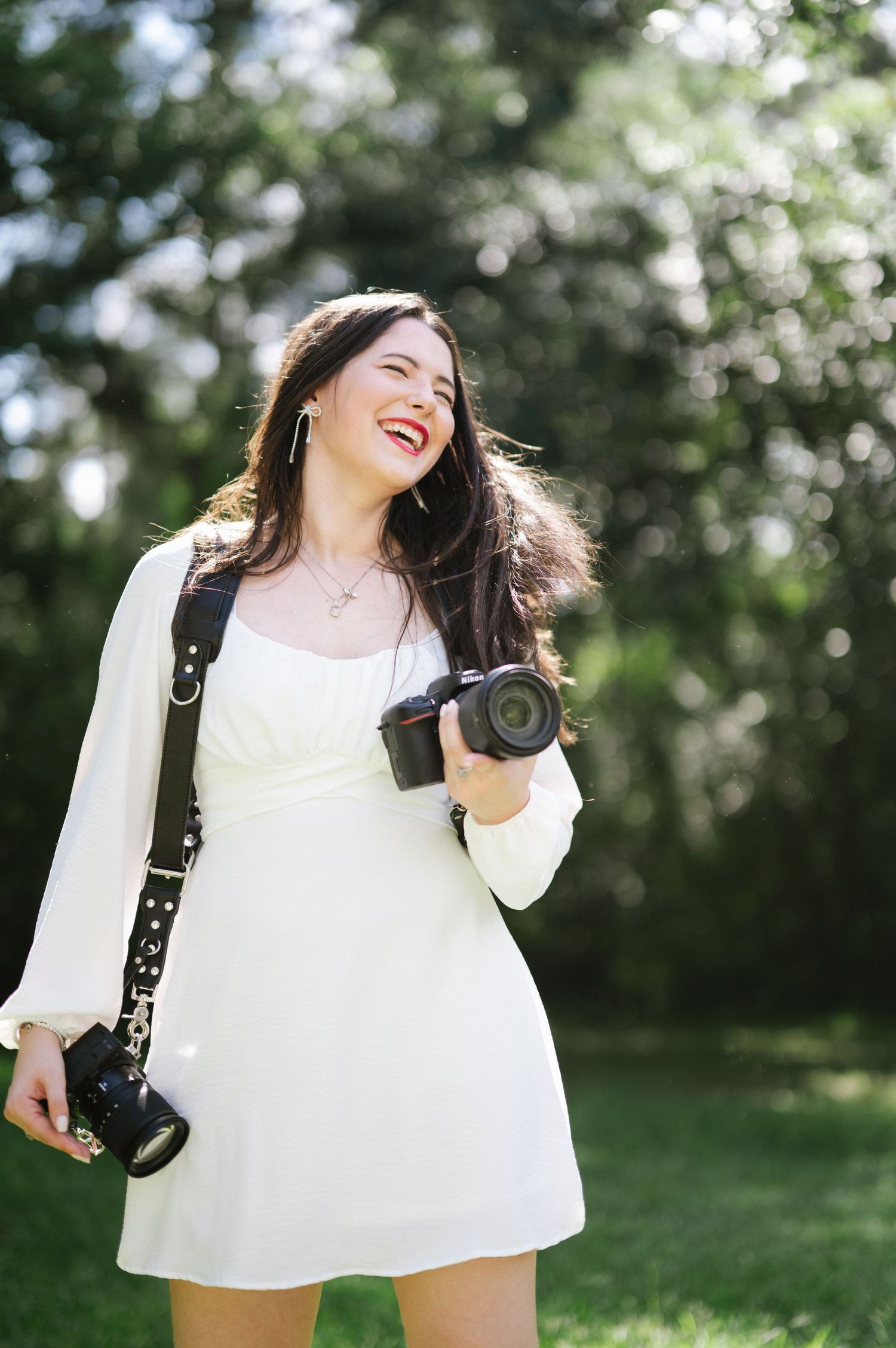 Happy female photographer in white dress laughing while holding camera equipment in sunny green park.