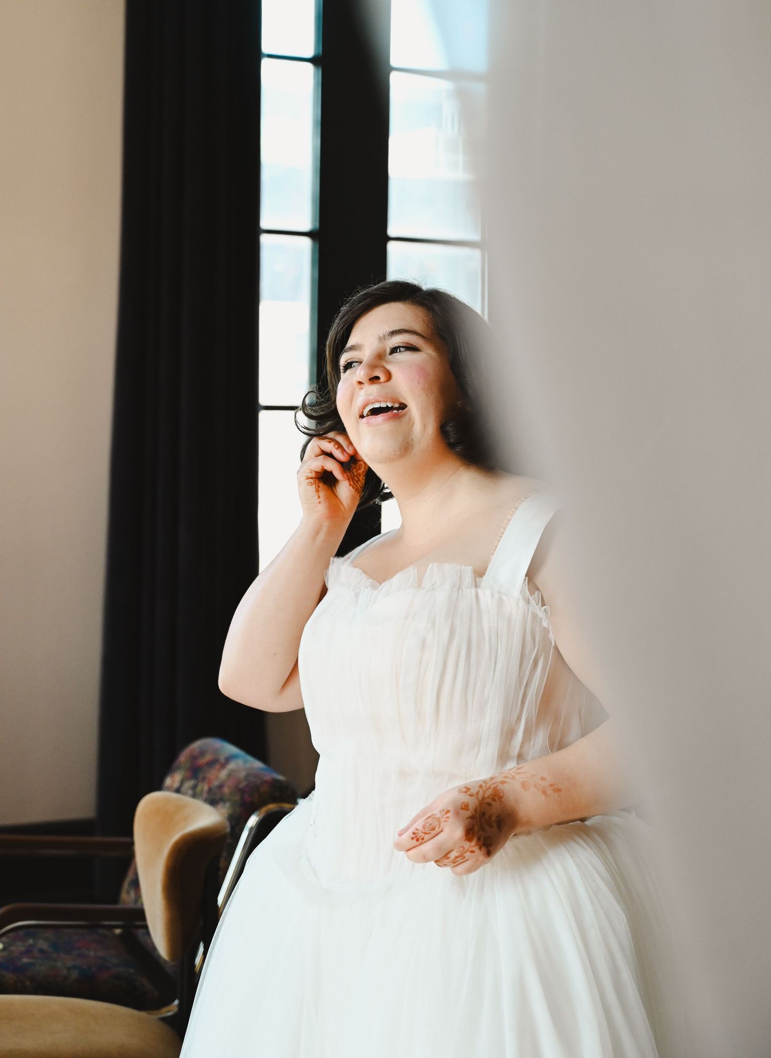 A joyful bride in a white tulle gown laughs while on the phone near a dark window in soft natural light.