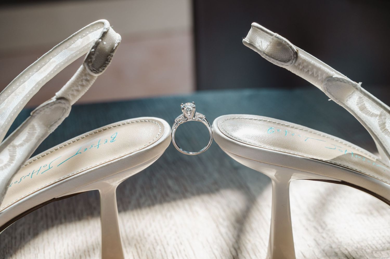 Diamond engagement ring nestled between white satin bridal heels on a teal surface, elegant wedding detail shot.