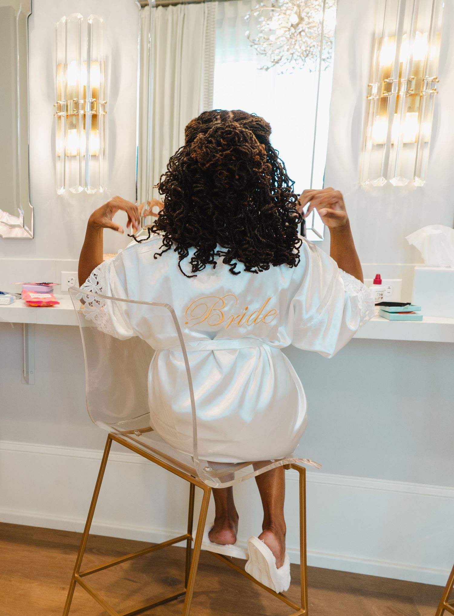 Bride in white satin robe sitting at vanity mirror getting ready, with curly hair and gold script lettering.