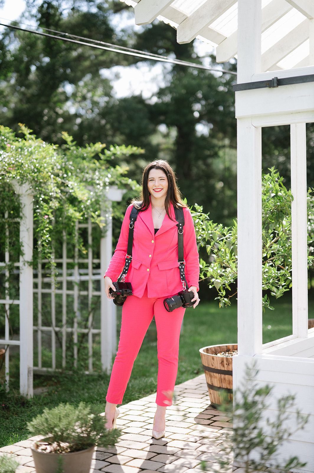 Woman in pink suit holding cameras smiles in a lush garden near a white pergola.