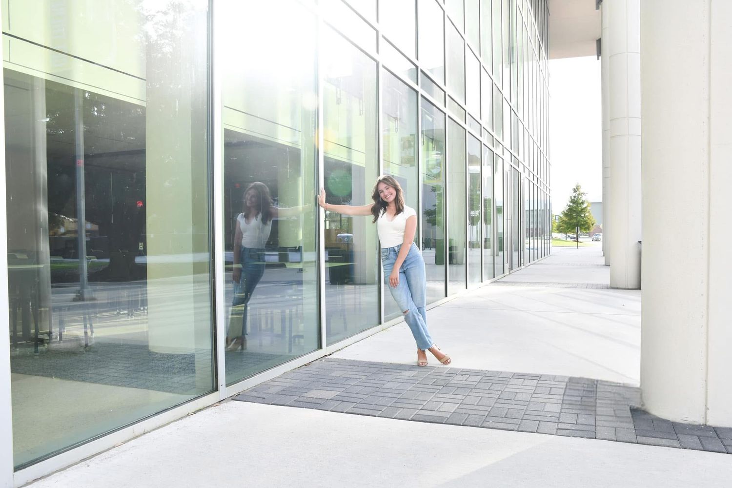Young woman in white top and jeans posing outside a modern glass building.