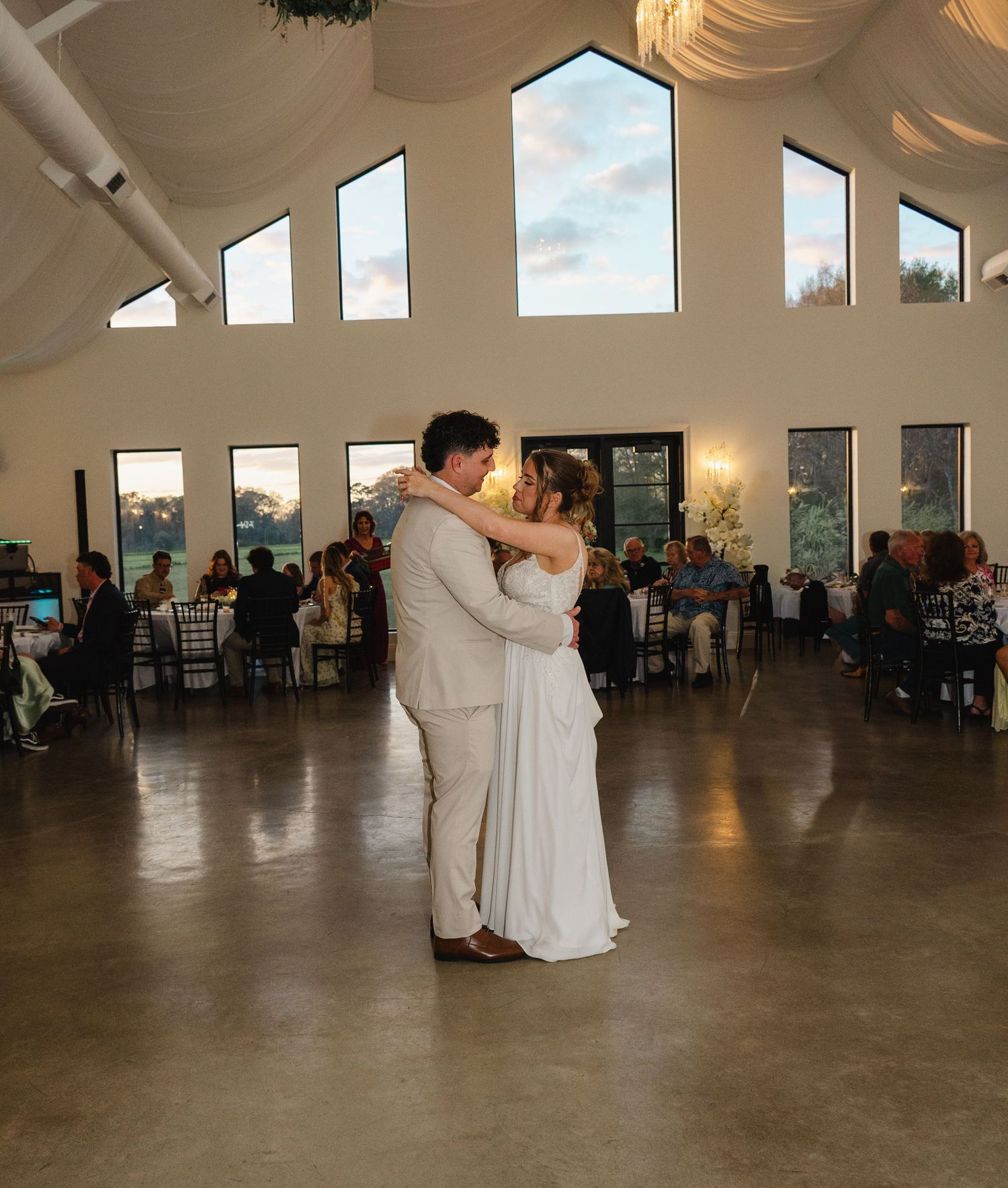Bride and groom share their first dance in a bright reception hall with vaulted ceilings and large windows.