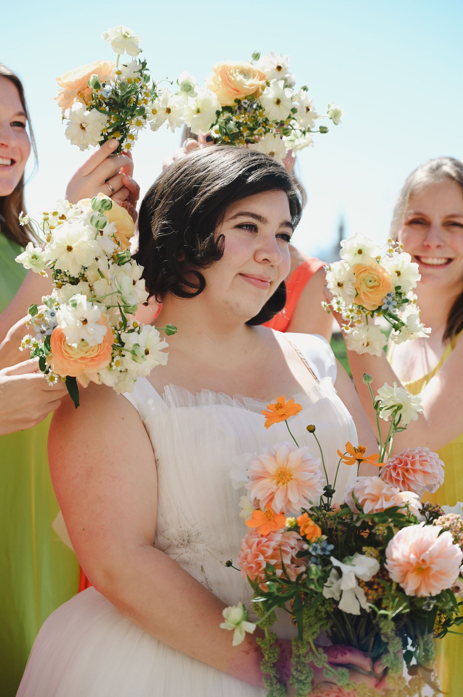 Bride holding peach and white floral bouquet surrounded by bridesmaids lifting bouquets overhead on sunny wedding day.