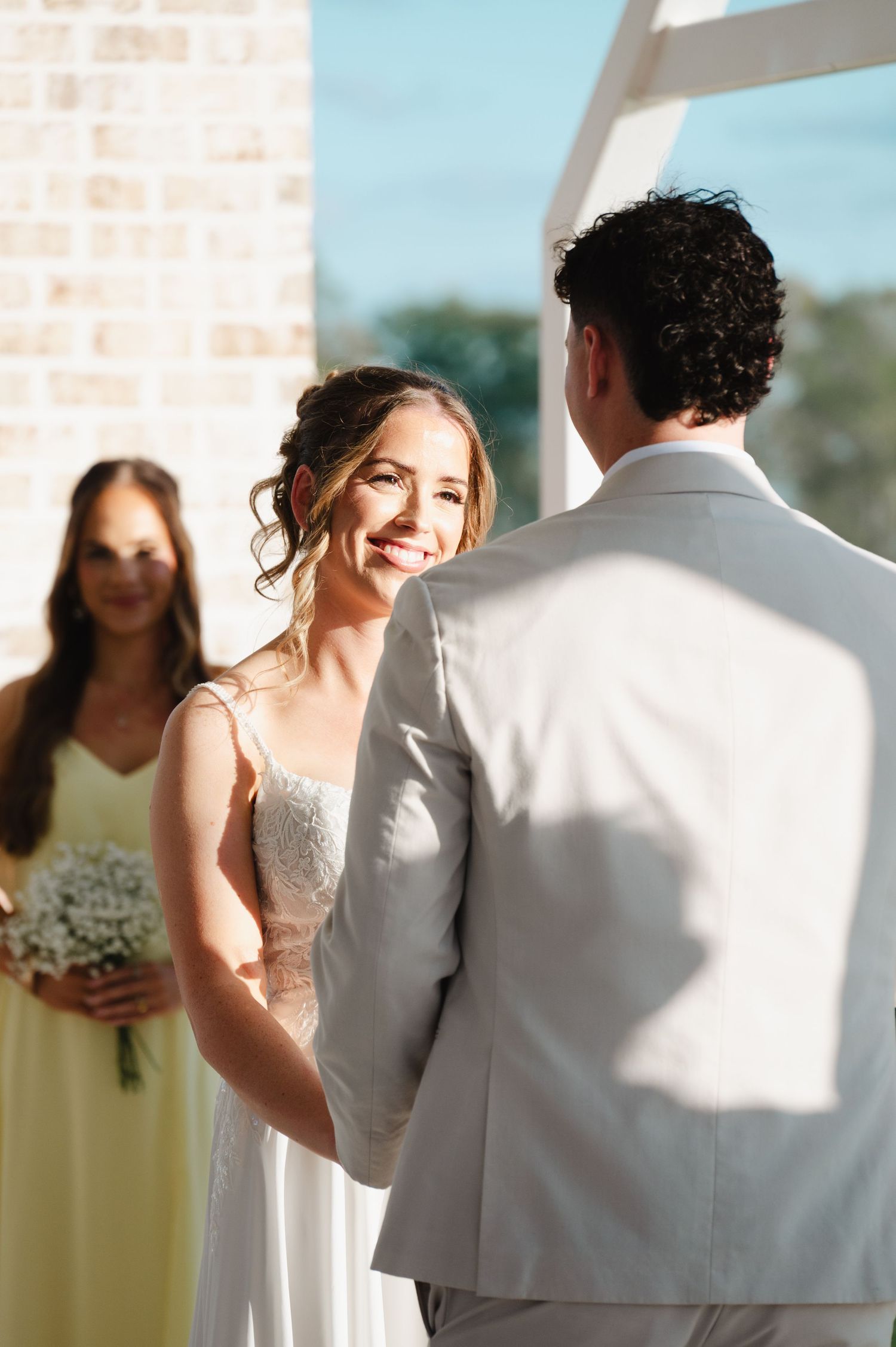 Bride smiles radiantly at groom during outdoor wedding ceremony while bridesmaid holds bouquet in background.