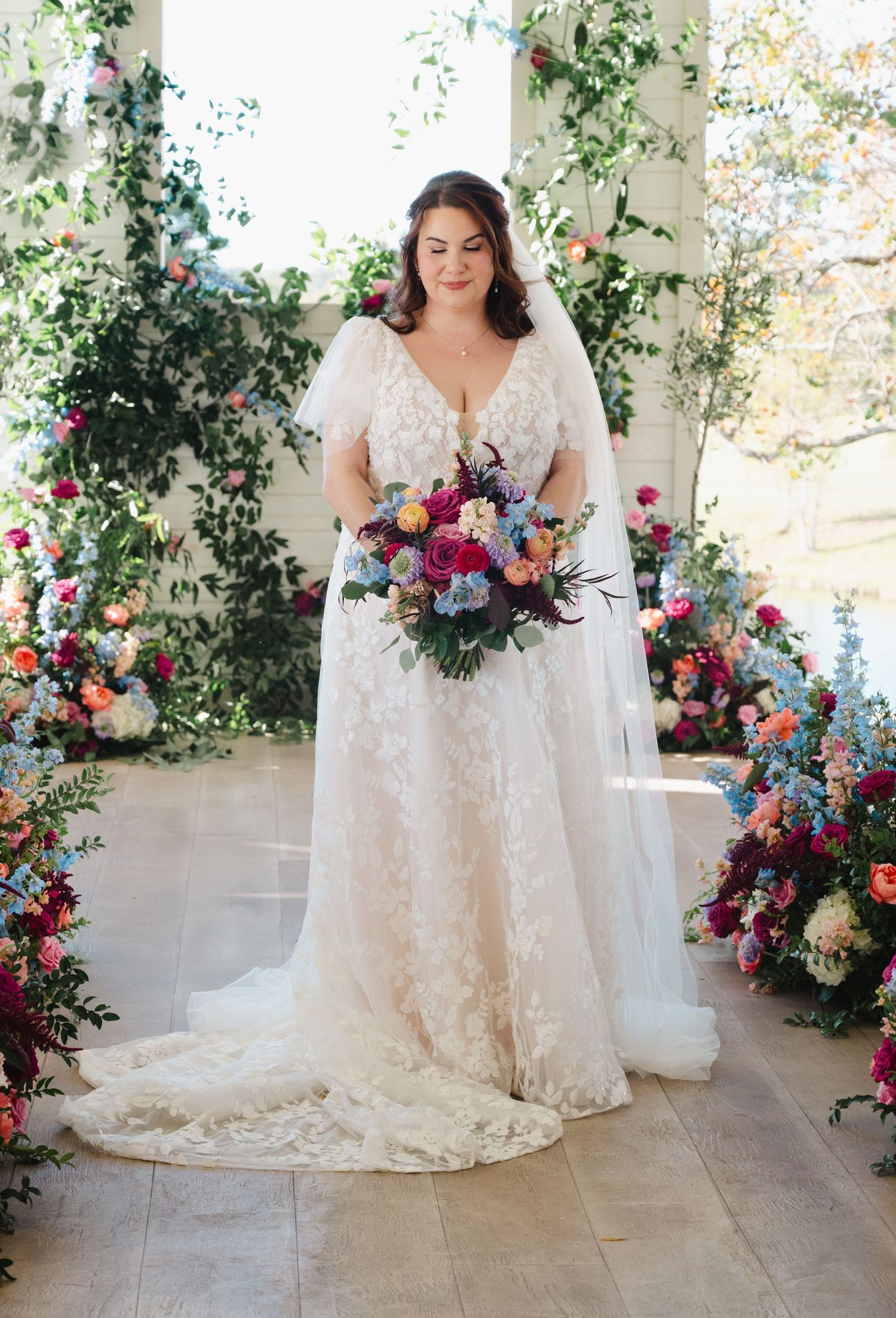 Bride in lace flutter-sleeve wedding gown holds colorful bouquet before vibrant floral arch backdrop.