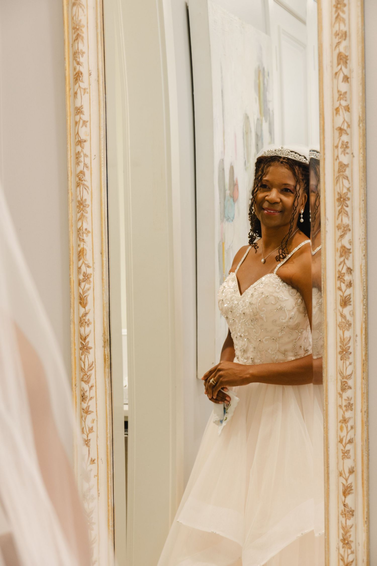 Smiling bride in beaded white gown and tiara admires her reflection in an ornate gold-framed mirror.