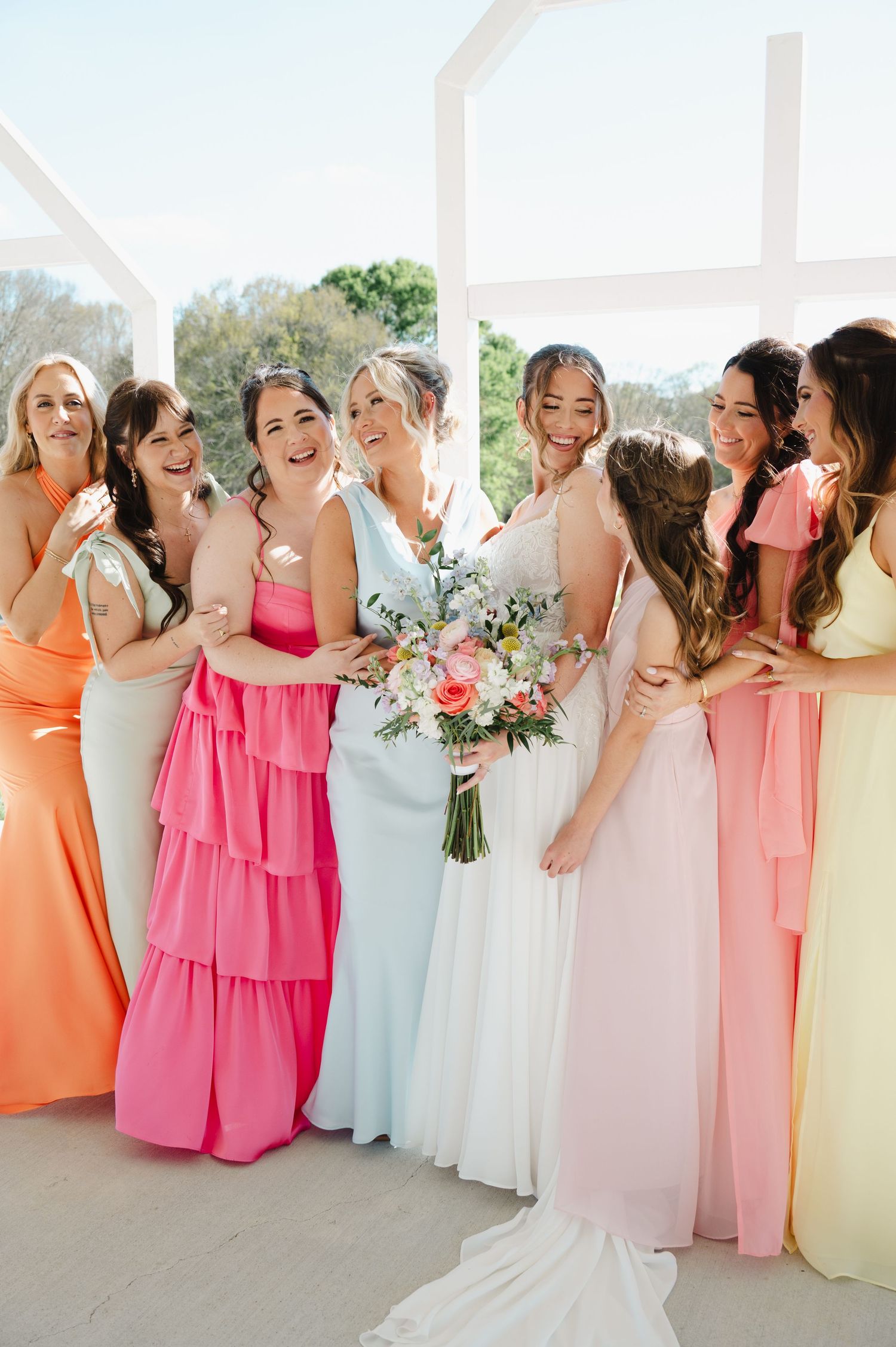 Bride in white gown poses with six bridesmaids in colorful pastel dresses holding floral bouquets.