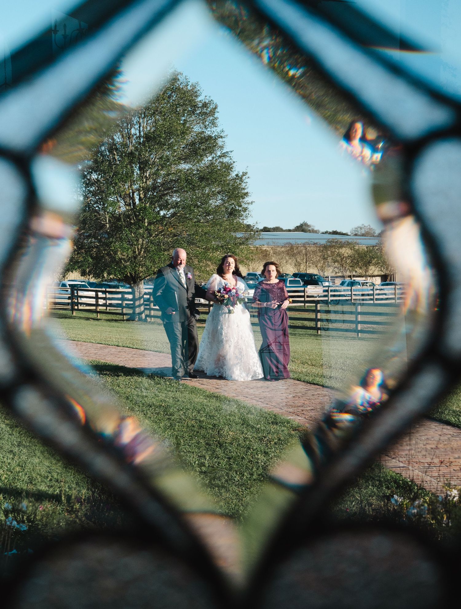Bride walking down outdoor path with two escorts at a farm wedding venue, photographed through decorative glass.