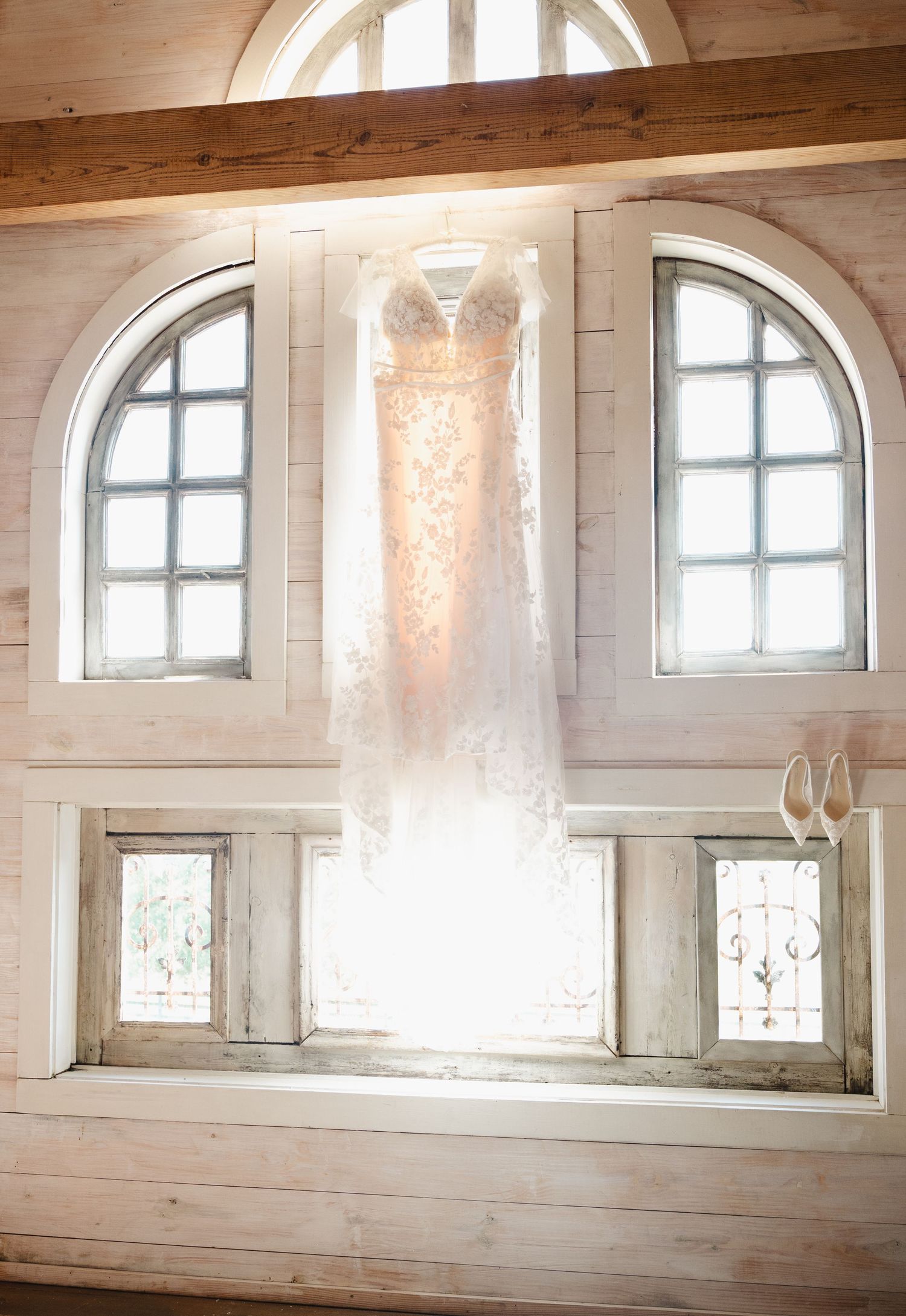 A wedding dress hanging in front of arched windows in a rustic white wood chapel, backlit by natural light.