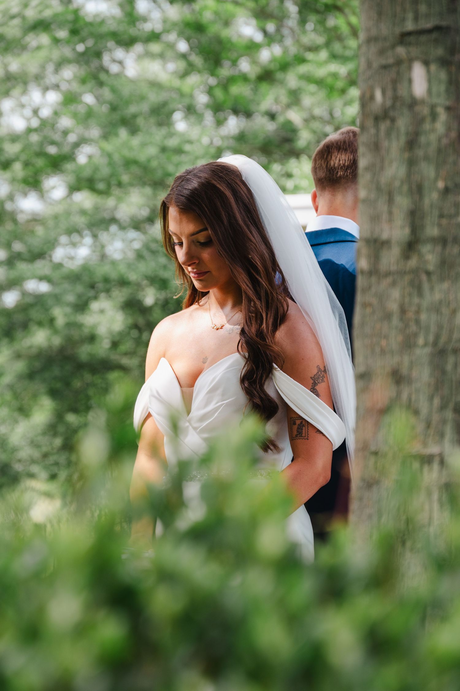 Bride in off-shoulder wedding gown and veil stands near a tree in lush greenery, groom in blue suit behind her.
