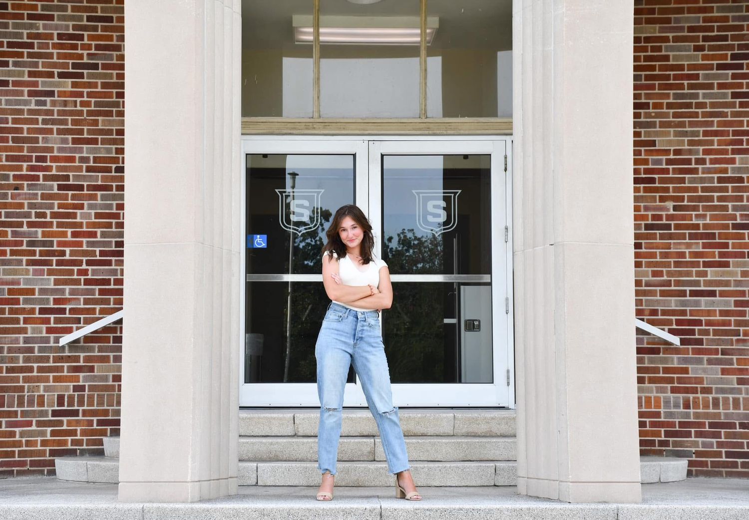 Young woman in white top and jeans standing confidently on steps of a brick building.