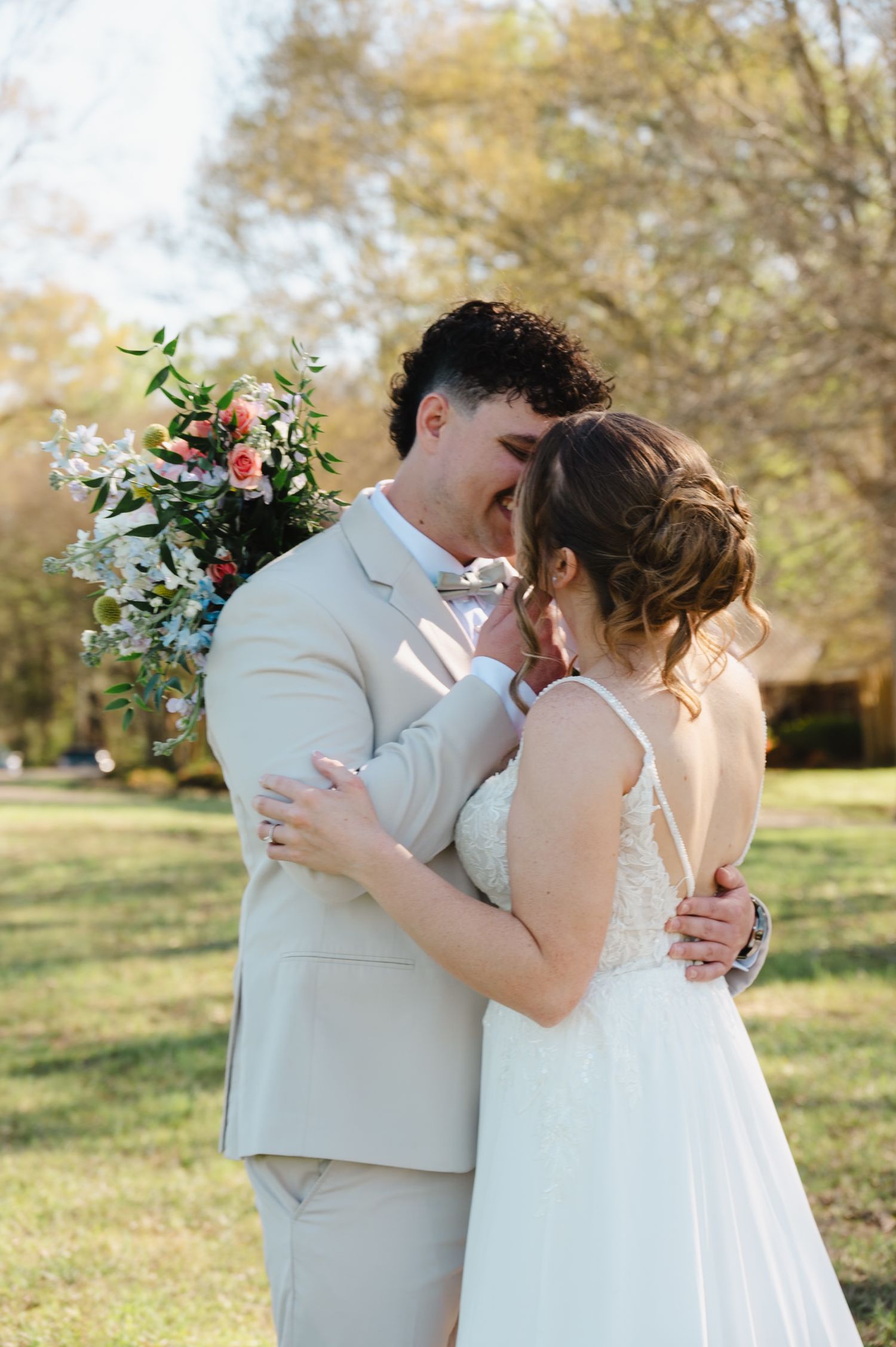 Groom in tan suit and bride in lace gown share intimate kiss outdoors holding floral bouquet.