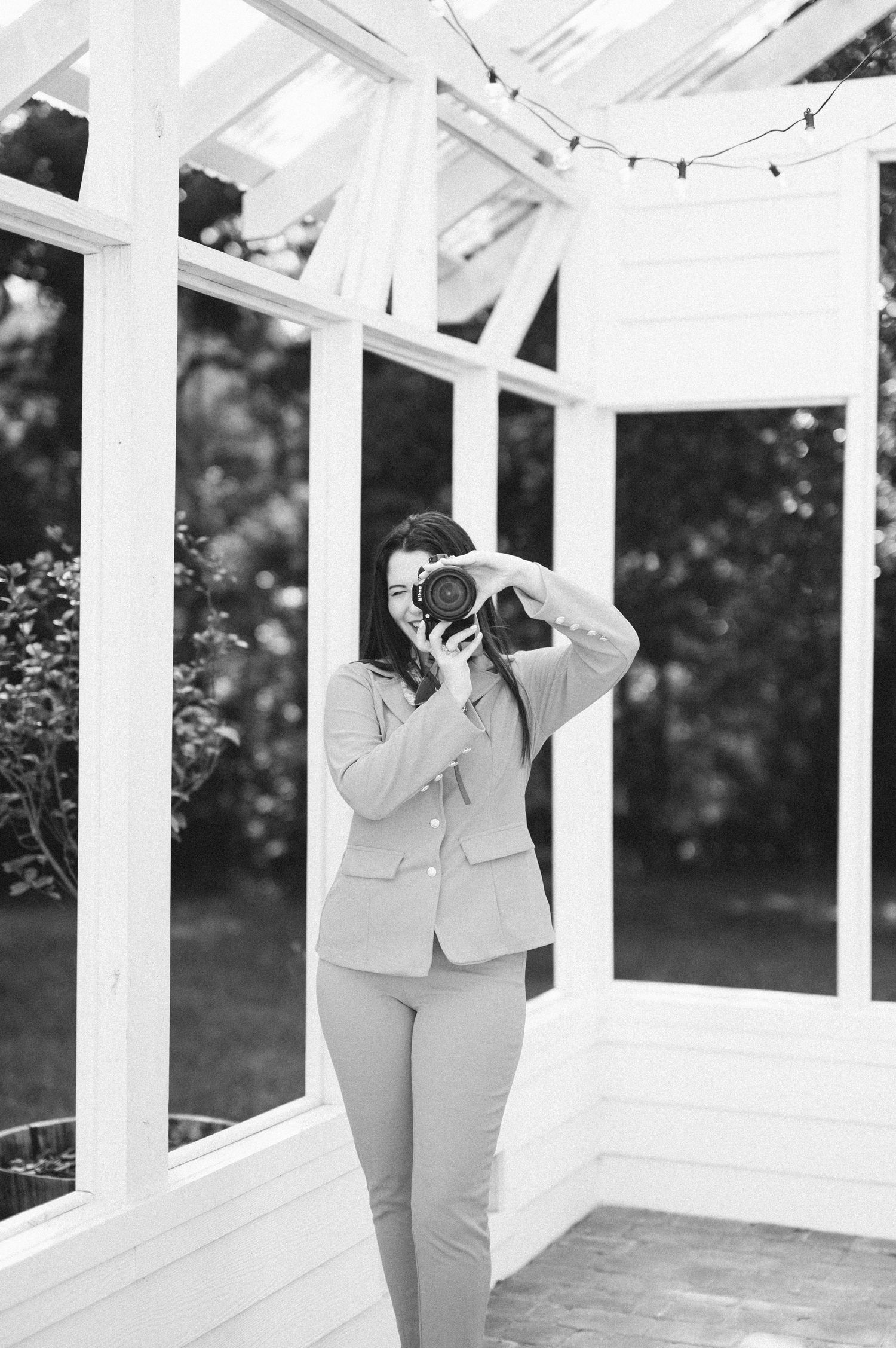 A female photographer holds a camera to her face inside a white greenhouse in black and white.
