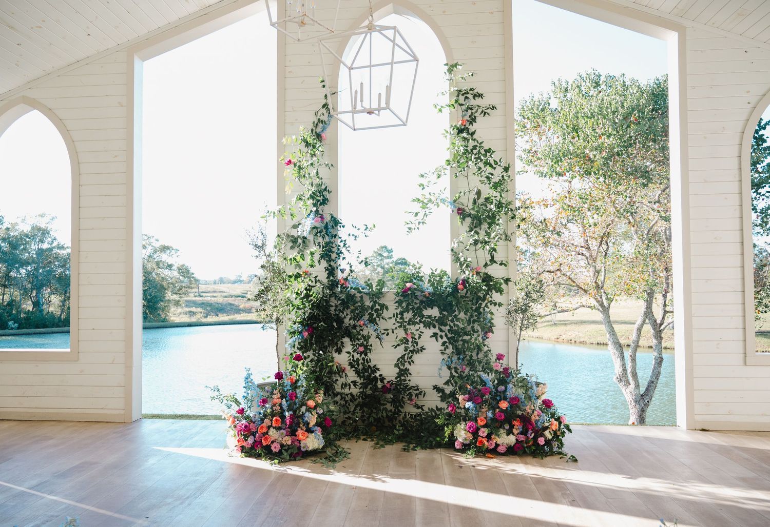 Elegant wedding ceremony backdrop with lush greenery and colorful floral arrangements beside arched windows overlooking a serene waterway.
