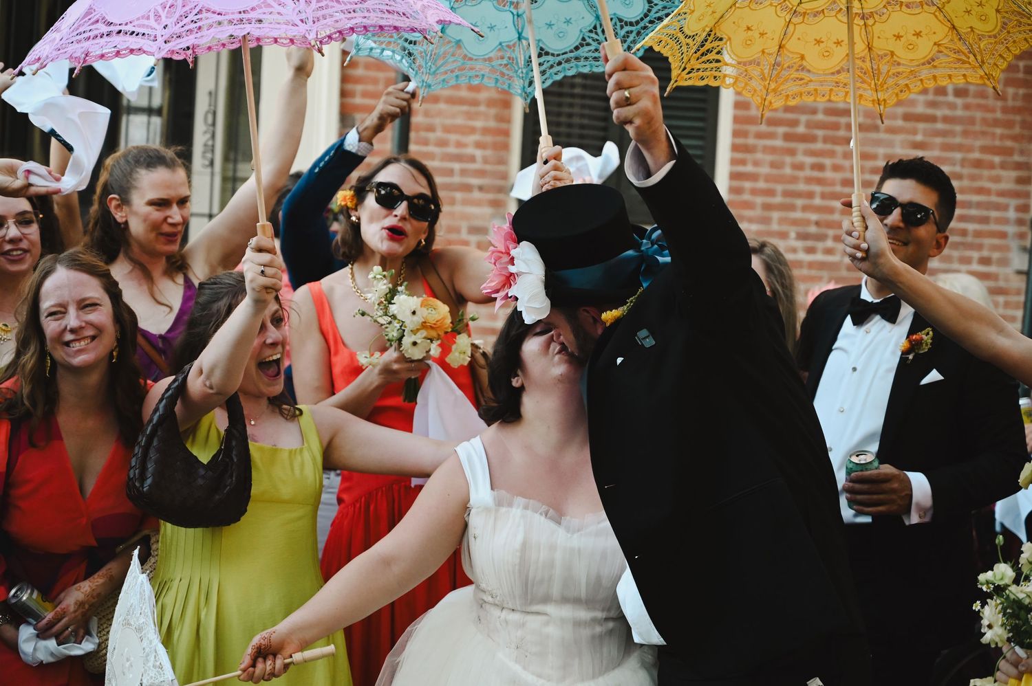 Bride and groom celebrate outdoors with guests holding colorful parasols at a festive wedding reception.