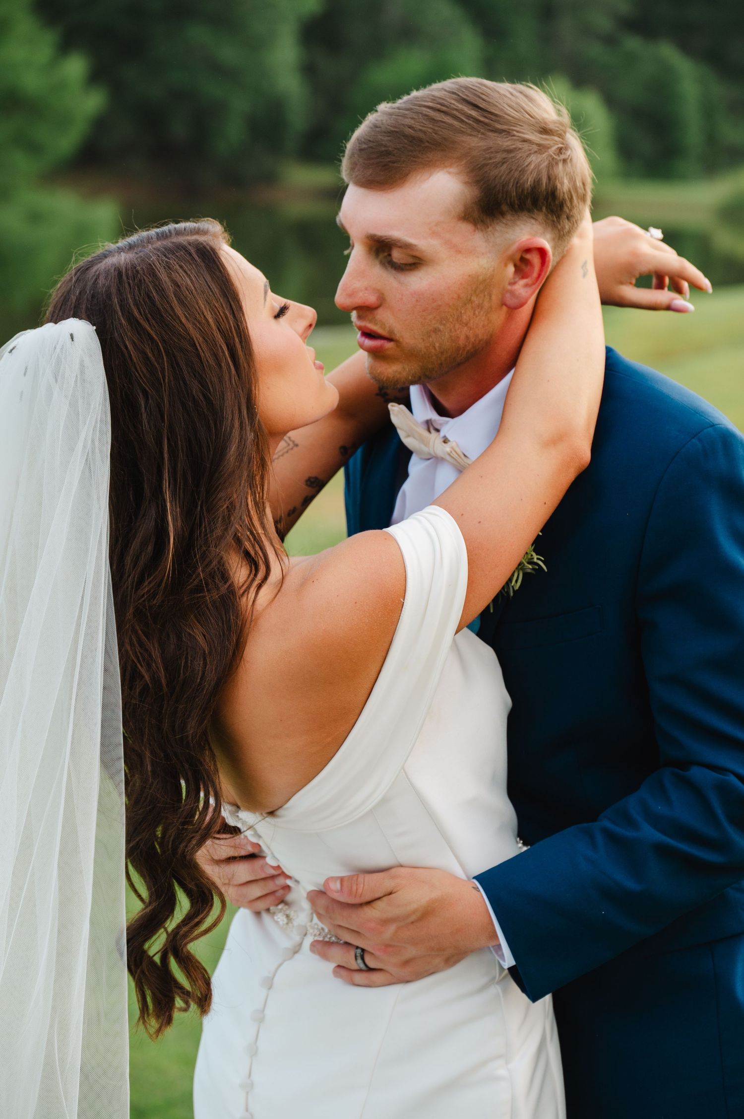 Bride in white gown and veil embraces groom in navy suit outdoors during romantic wedding portrait session.