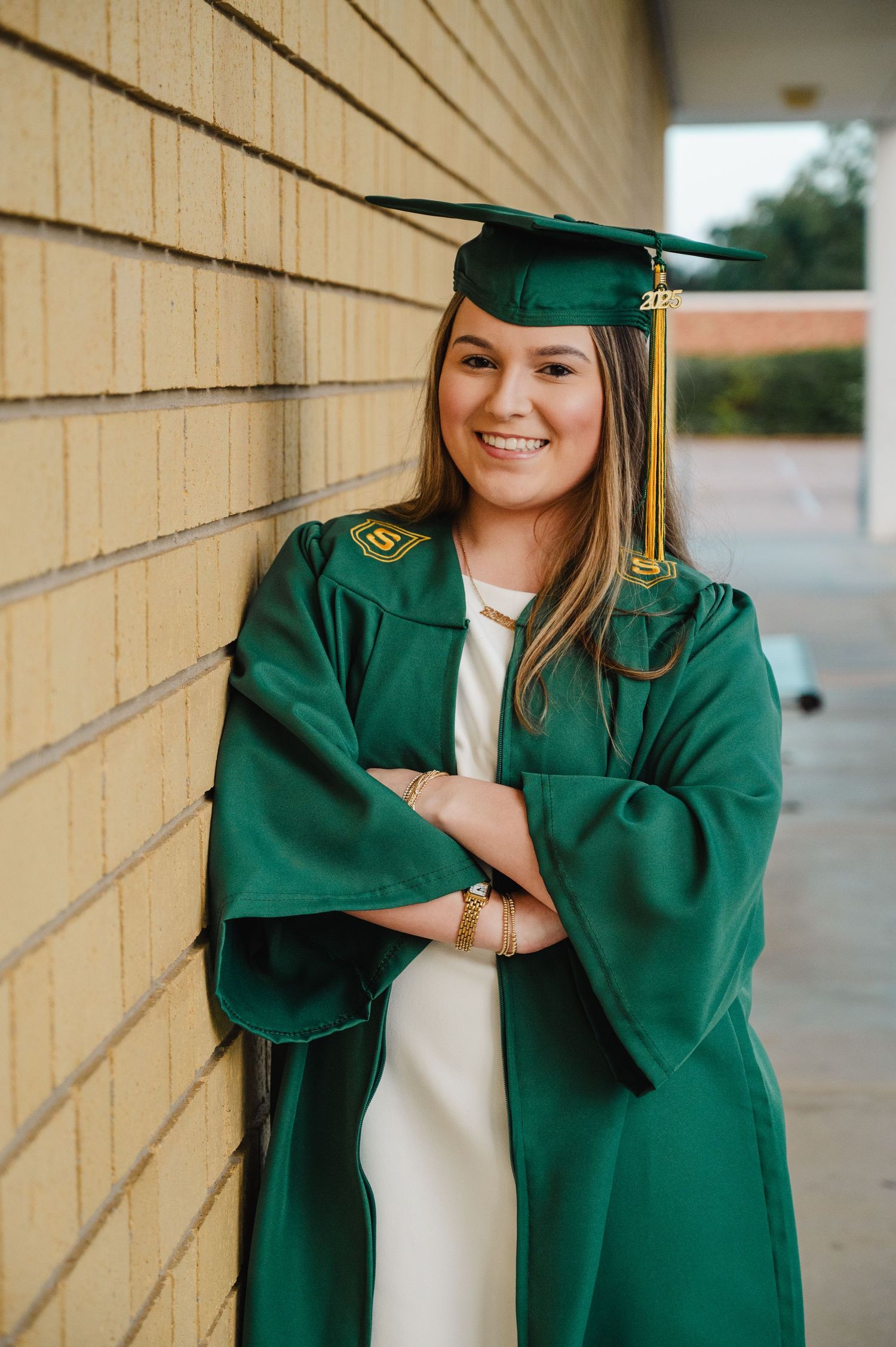 Smiling graduate in green cap and gown leans against brick wall with arms crossed during outdoor graduation photo.
