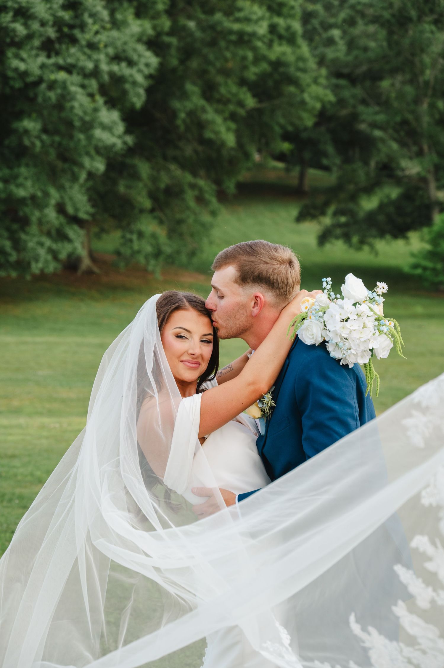 Bride and groom share a romantic kiss outdoors, her white veil flowing, as she holds a bouquet of white flowers.
