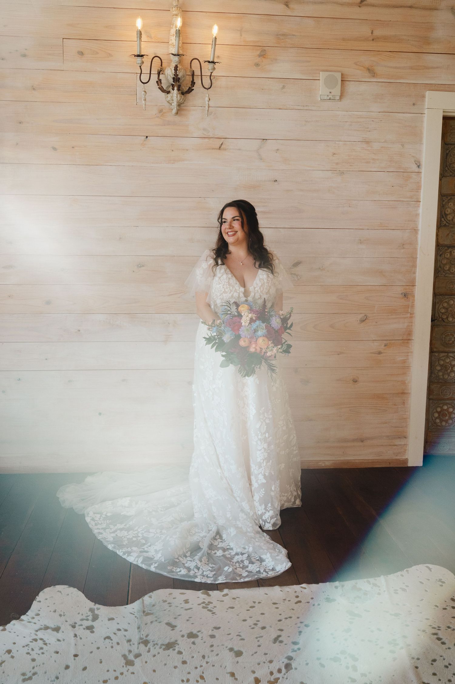 Bride in lace wedding gown holding floral bouquet, smiling in rustic wood-paneled room with chandelier.