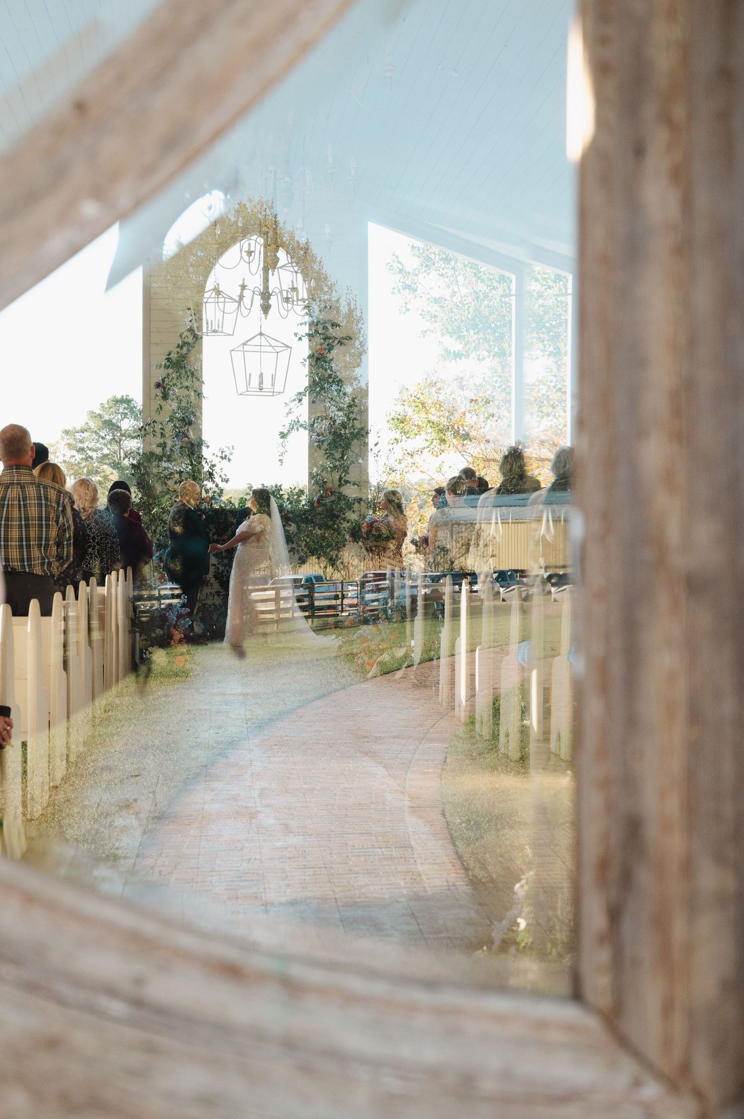 A bride walks down a rustic aisle framed by white pews and floral arch, viewed through a weathered wooden window frame.