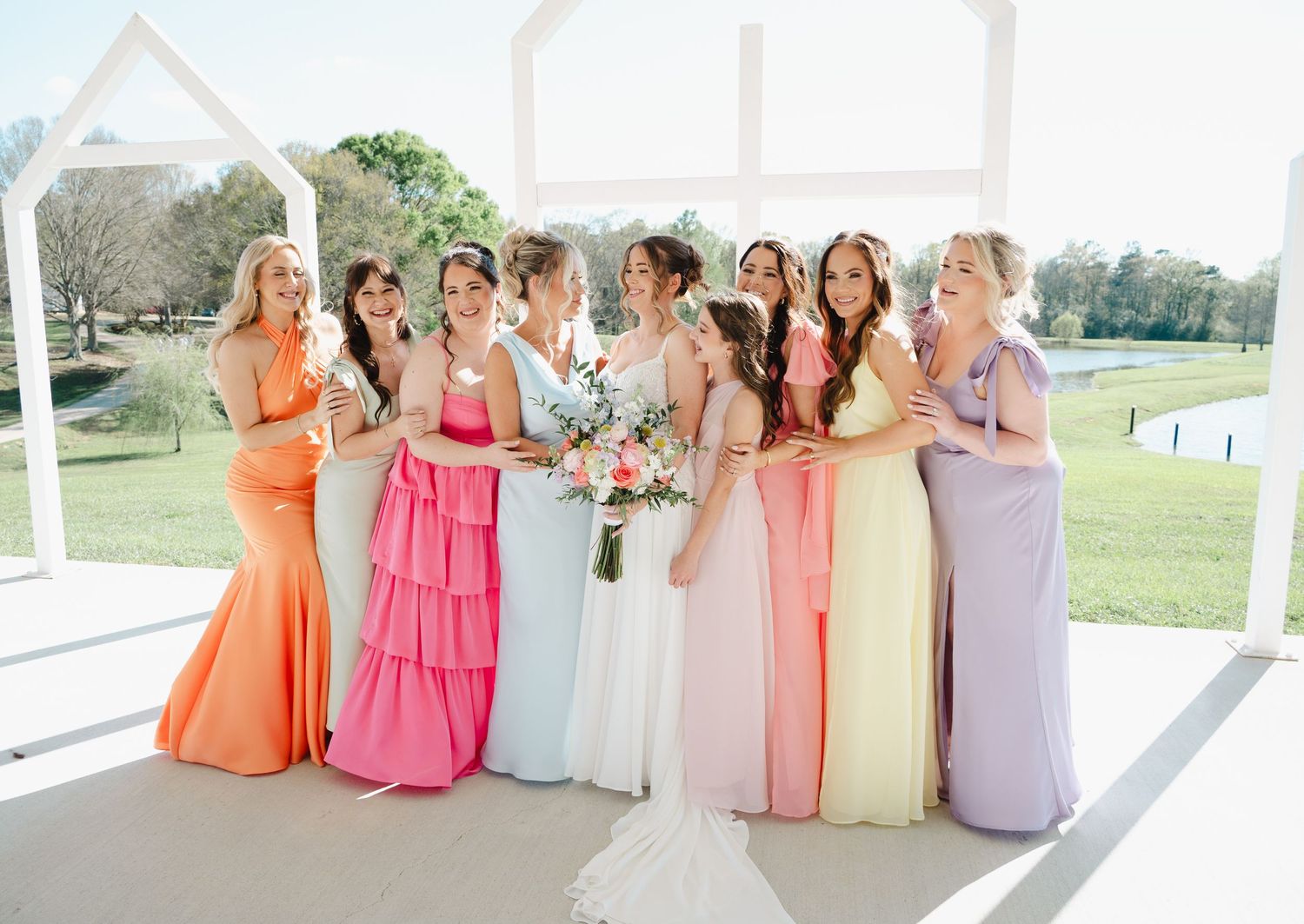 Bride with bridesmaids in pastel dresses laugh together under a white wedding arch on a sunny day.