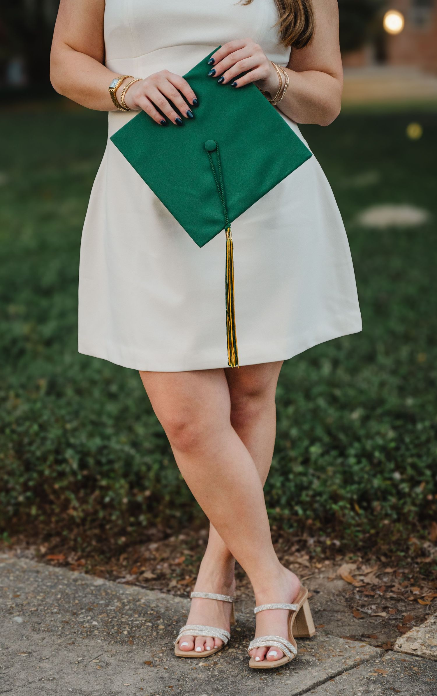 Graduate in white dress holding green cap with gold tassel, wearing heeled sandals outdoors.