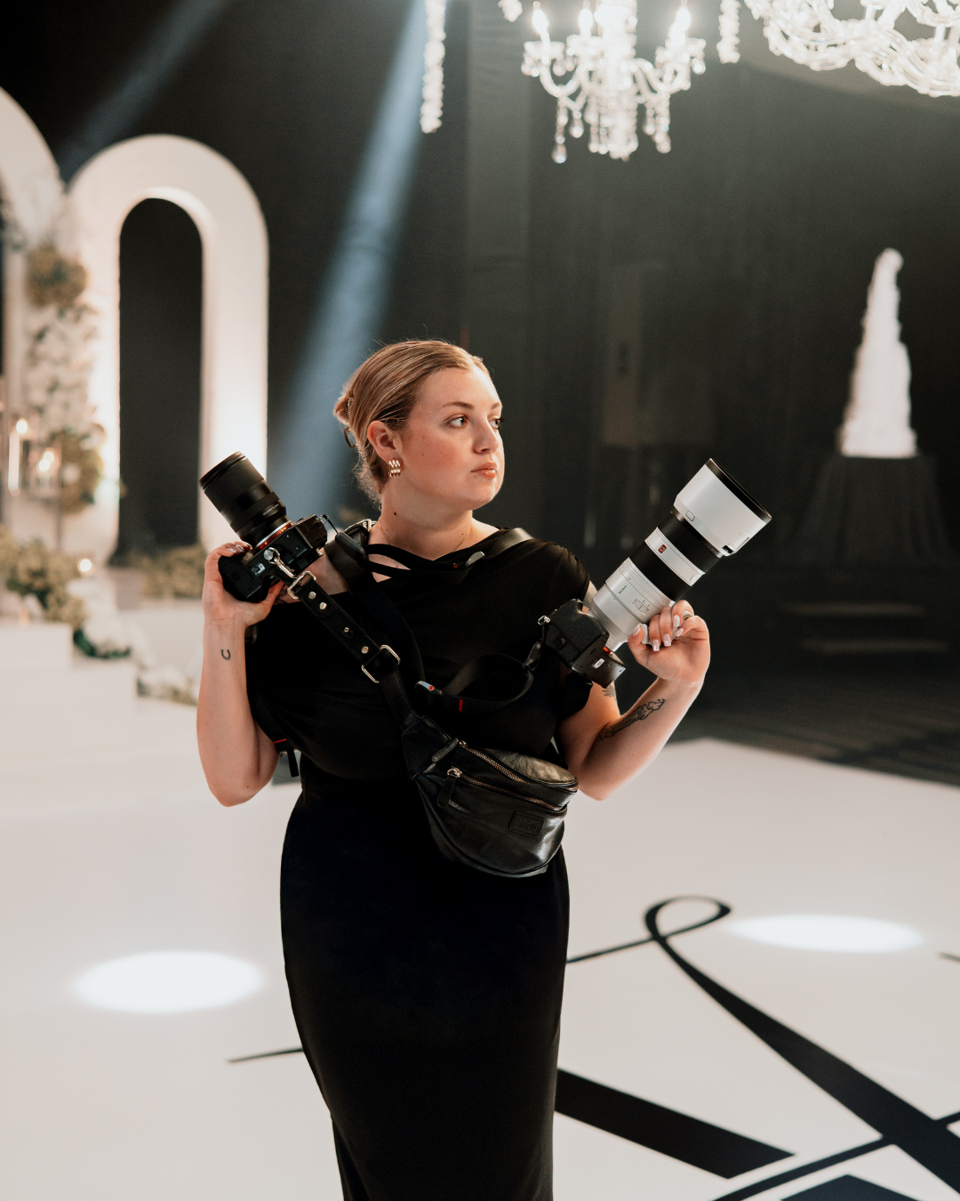 A female photographer in a black dress holds two cameras at an elegant event venue with chandeliers and floral decor.