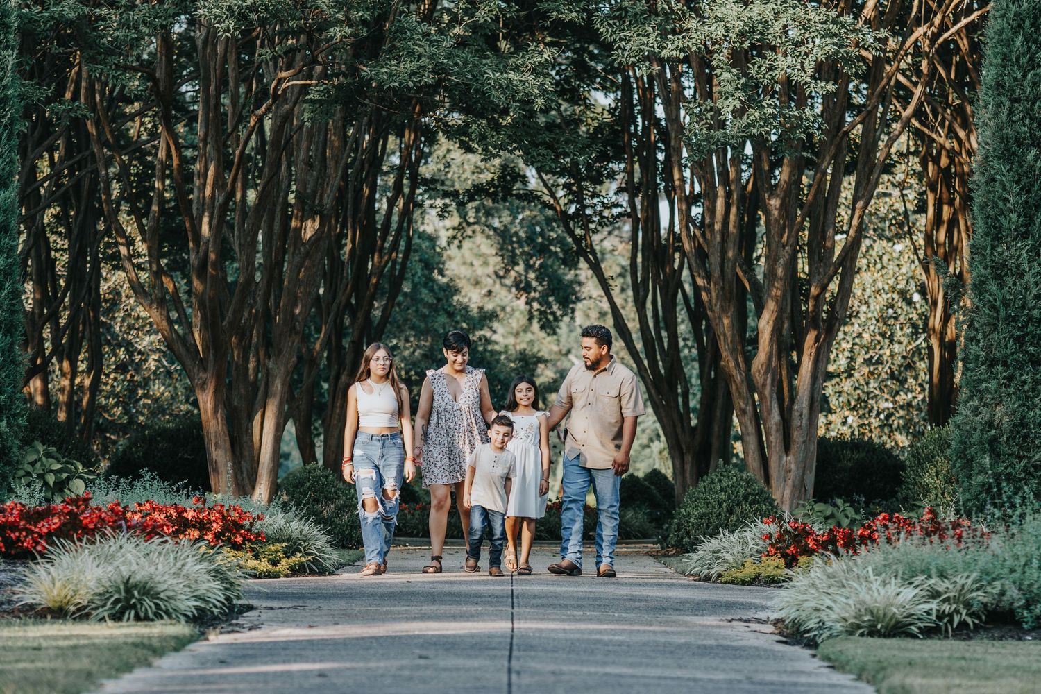 A group walks together on a tree-lined path with vibrant red flowers blooming along the edges on a sunny day.