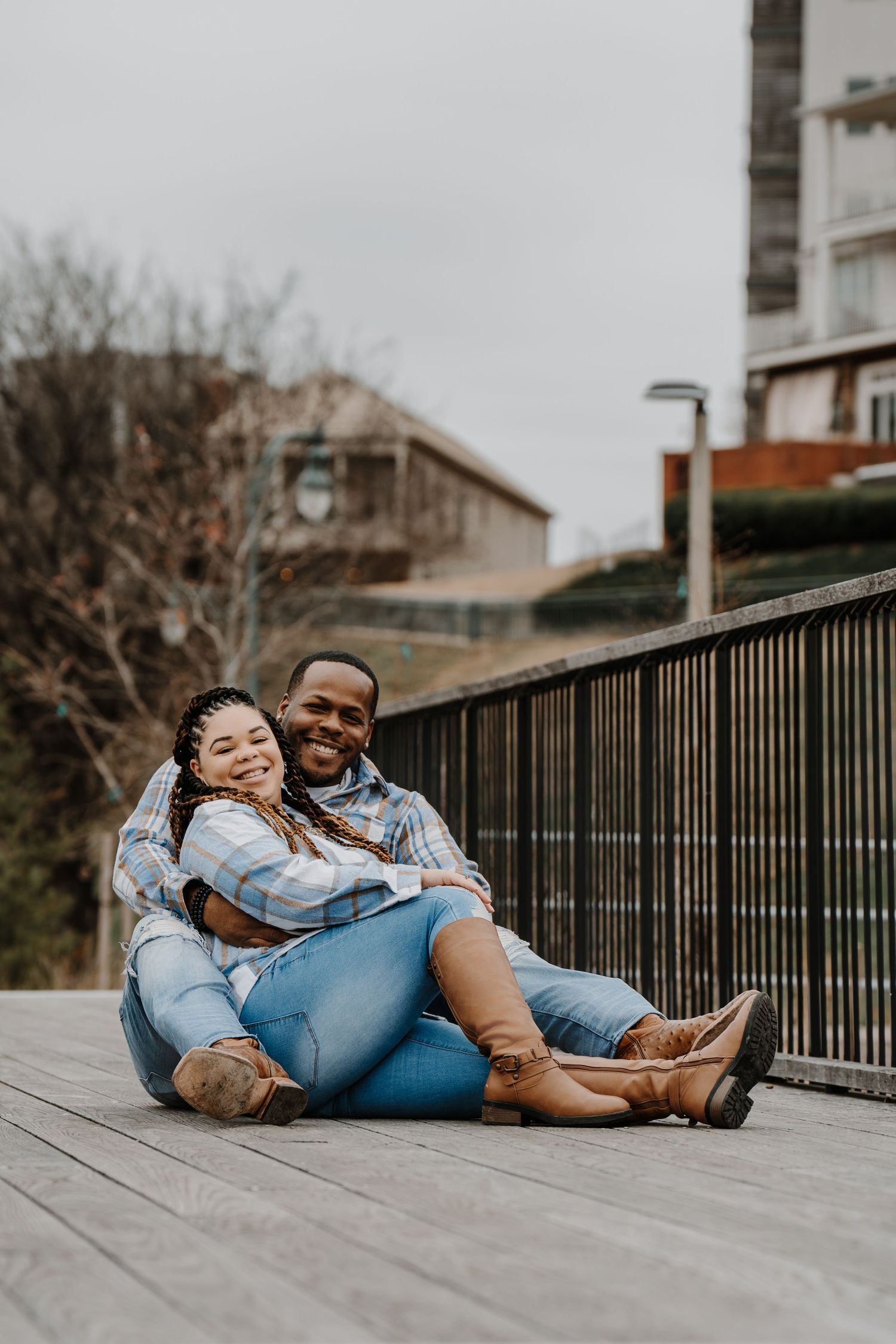 A couple in denim sits together on a wooden deck with metal railings on an overcast day.