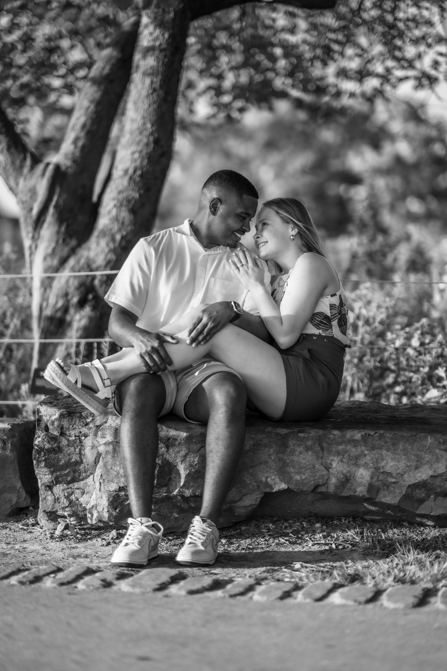A romantic black and white photograph of a couple sitting intimately together on stone steps under trees.
