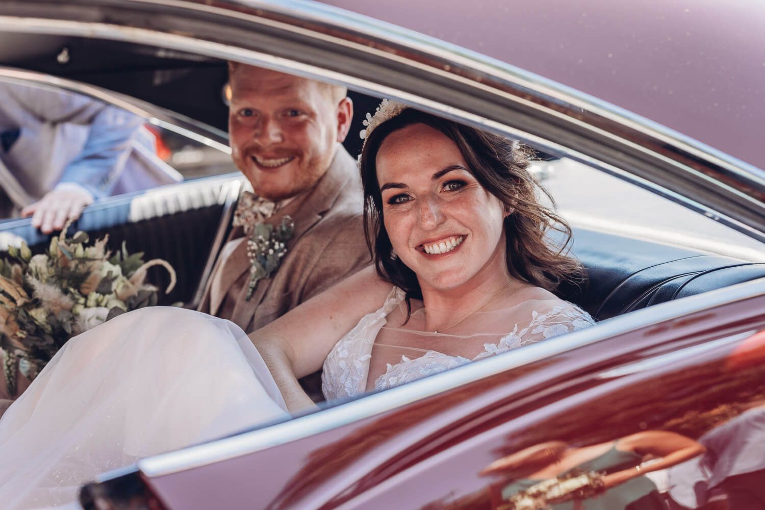 Glückliches Brautpaar sitzt nach der Hochzeit gemeinsam in einem Oldtimer und lächelt in die Kamera