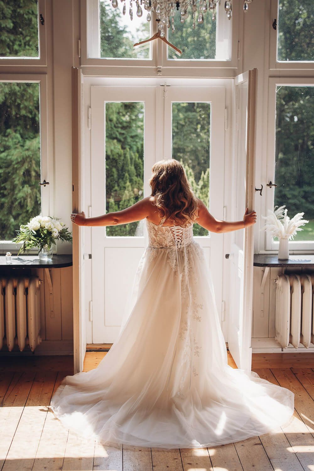 Braut steht im eleganten Hochzeitskleid vor geöffneten Türen im warmen Tageslicht, fotografiert von einer Fotografin in Hannover
