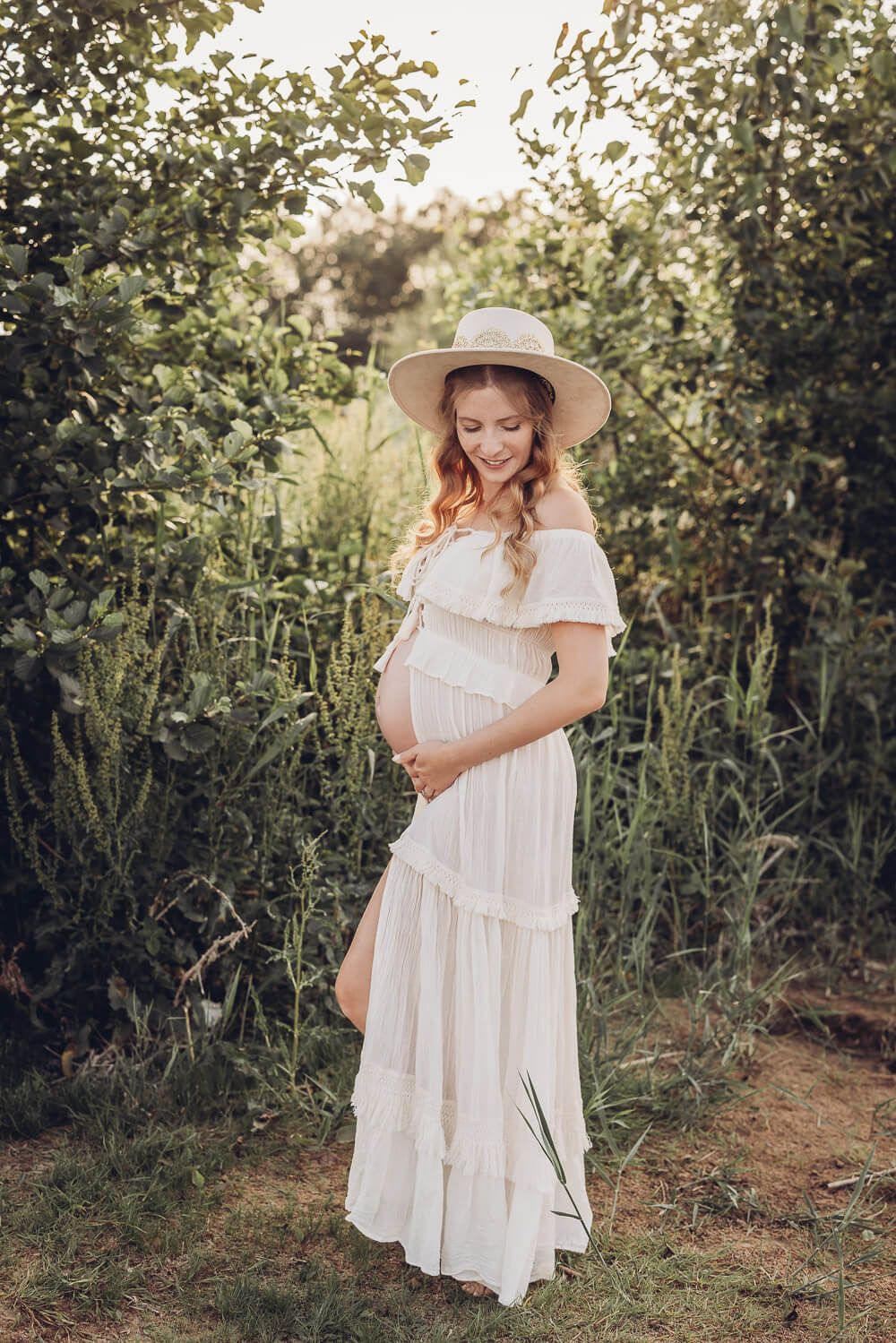 Schwangere Frau mit Hut steht in einer grünen Umgebung und hält ihren Babybauch - Fotoshooting in Celle