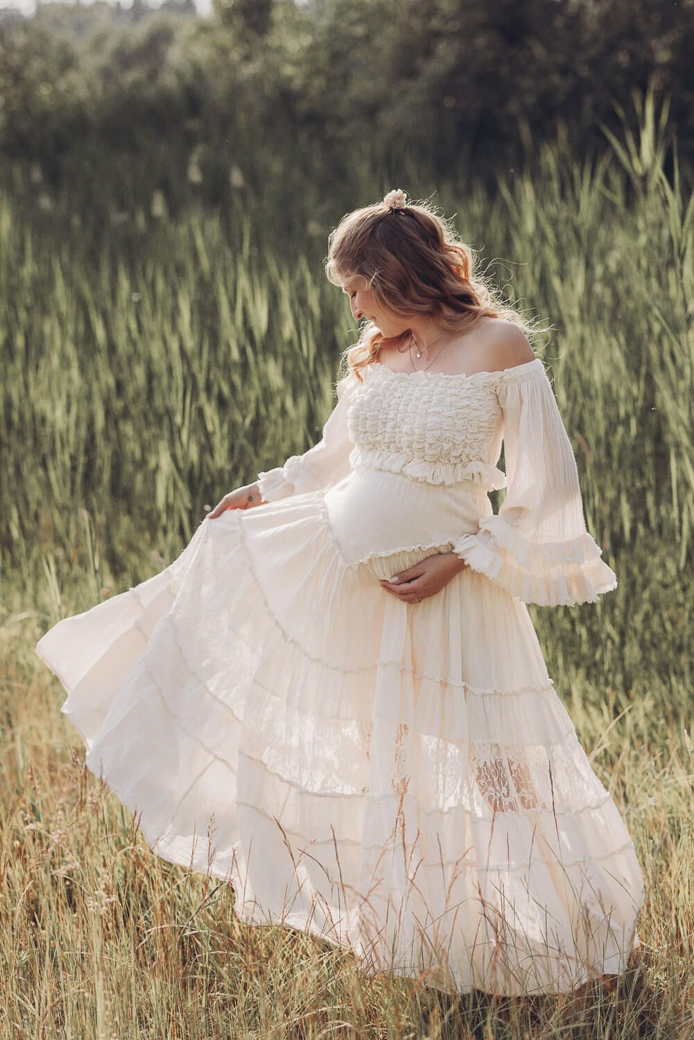Schwangere Frau in fließendem Boho-Kleid steht in einer Wiese und betrachtet ihren Babybauch
