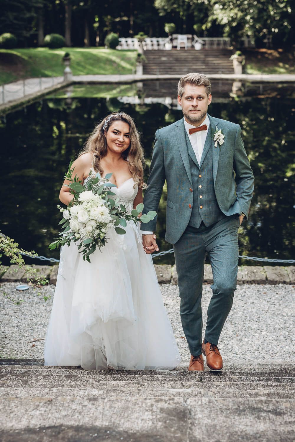 Brautpaar geht Hand in Hand an einem Teich entlang und blickt in die Kamera, aufgenommen von einem Hochzeitsfotografen in Braunschweig