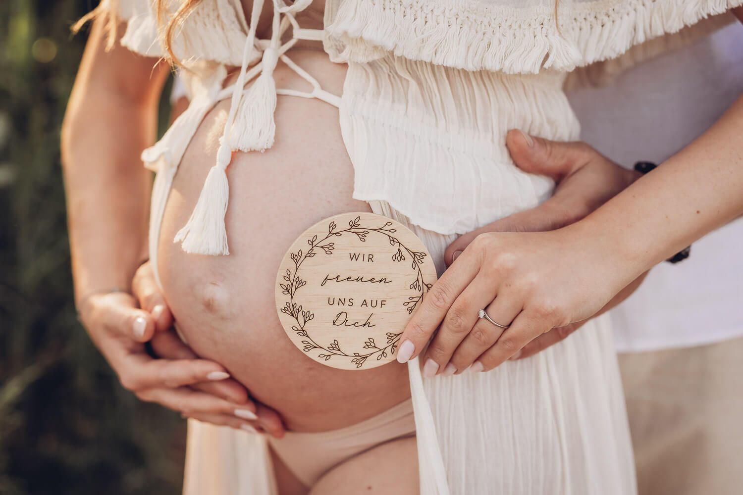 Detailaufnahme von Händen eines Paares auf einem Babybauch mit Holzschild und liebevoller Botschaft