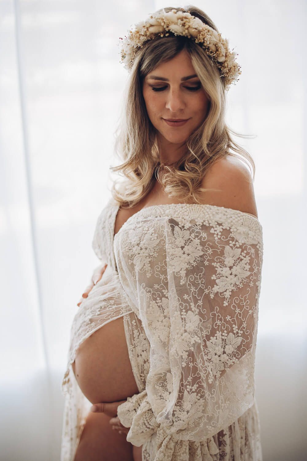 Stilvolles Babybauch Fotoshooting im Studio in Hannover – schwangere Frau mit Spitzenkleid und Blumenkranz betrachtet ihren Bauch