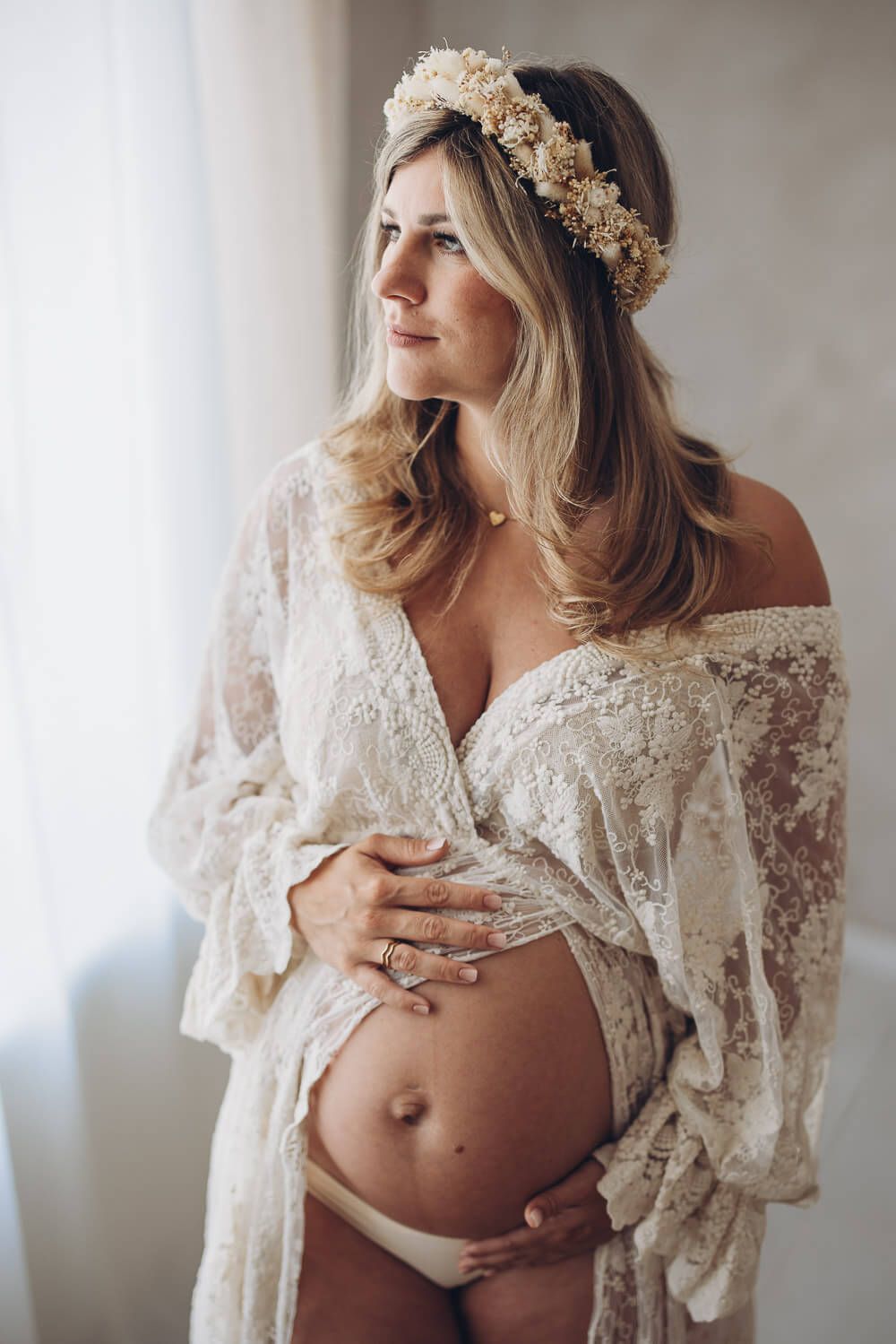 Elegantes Babybauch Fotoshooting im Fotostudio Gehrden – werdende Mutter mit Blumenkranz blickt ruhig aus dem Fenster