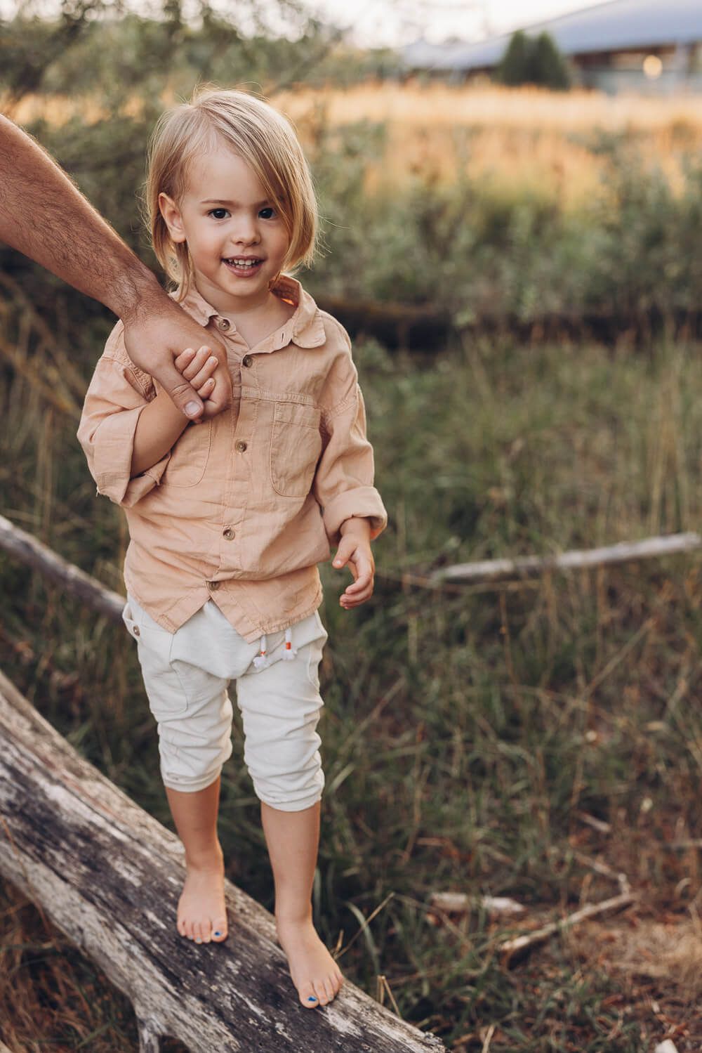 Natürliches Kinderportrait beim Outdoor Shooting Hannover, kleiner Junge steht auf einem Baumstamm und hält die Hand seines Vaters