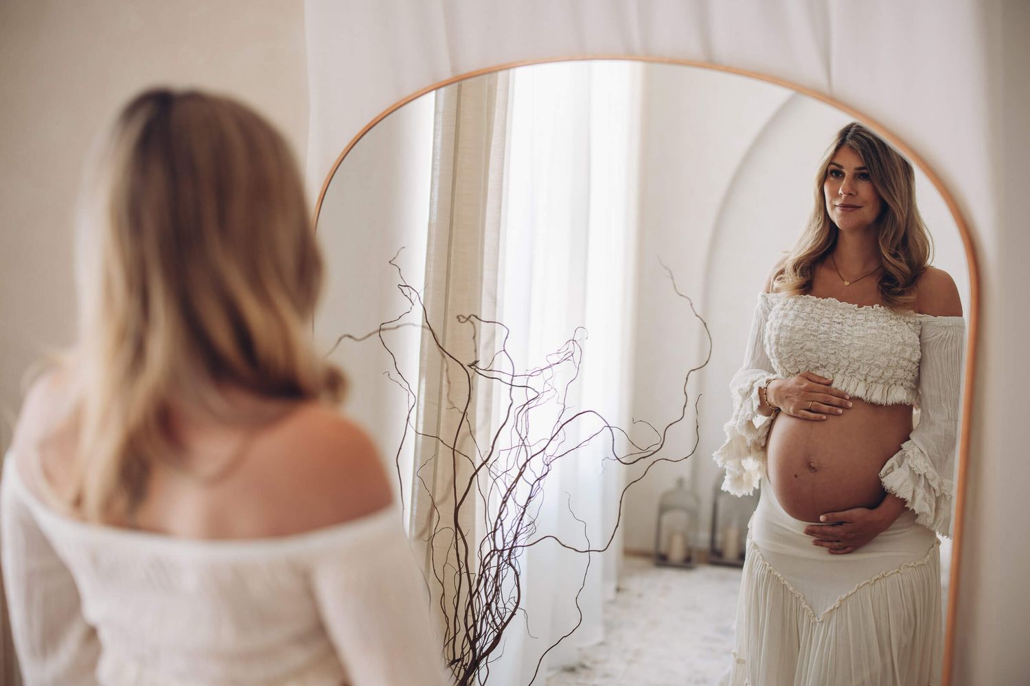 Schwangere Frau betrachtet liebevoll ihren Babybauch im Spiegel – ästhetisches Schwangerschafts-Fotoshooting im natürlichen Licht
