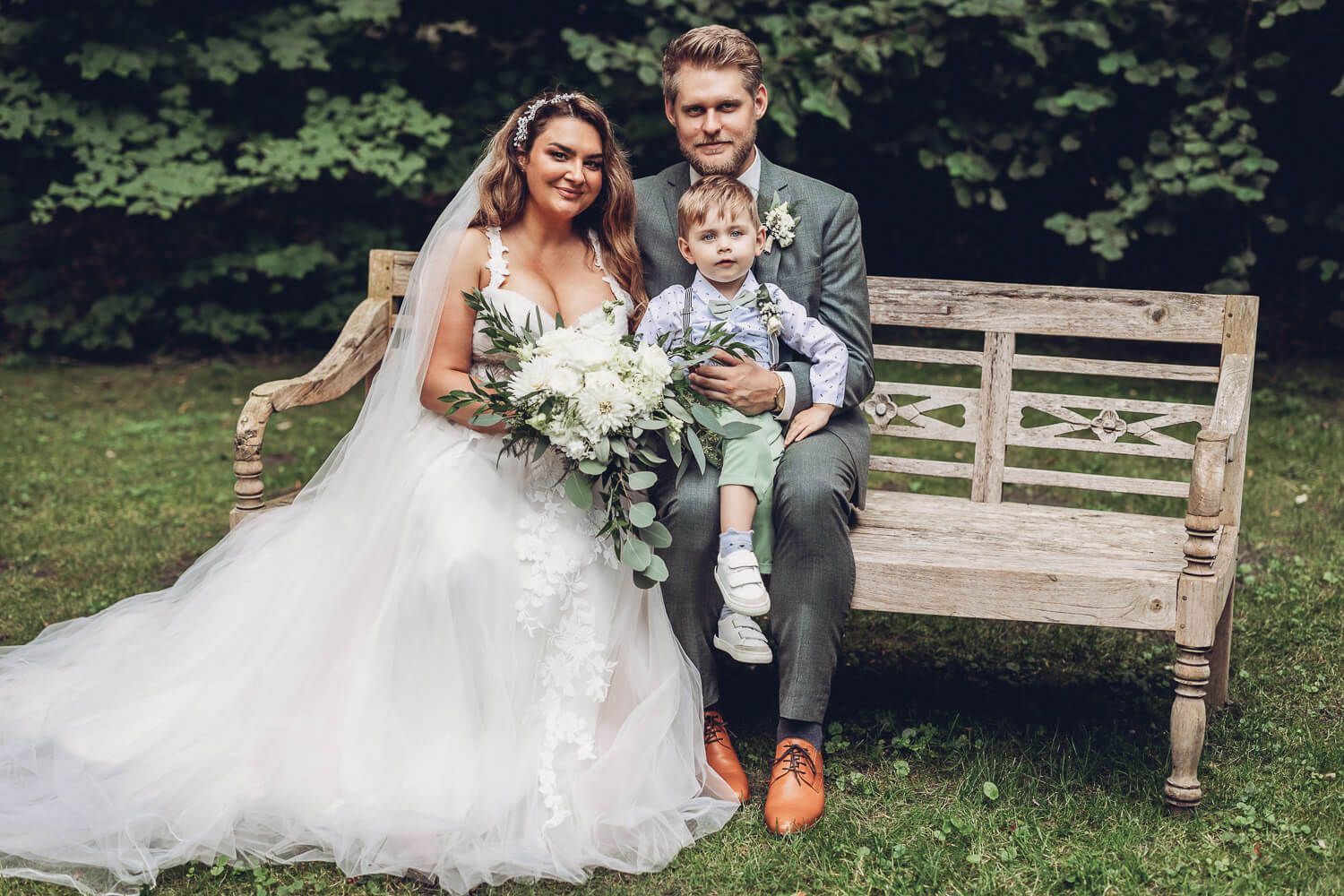 Brautpaar mit Kind sitzt gemeinsam auf einer Holzbank für ein natürliches Hochzeitsfoto im Grünen - Hochzeitsfotografin Hameln