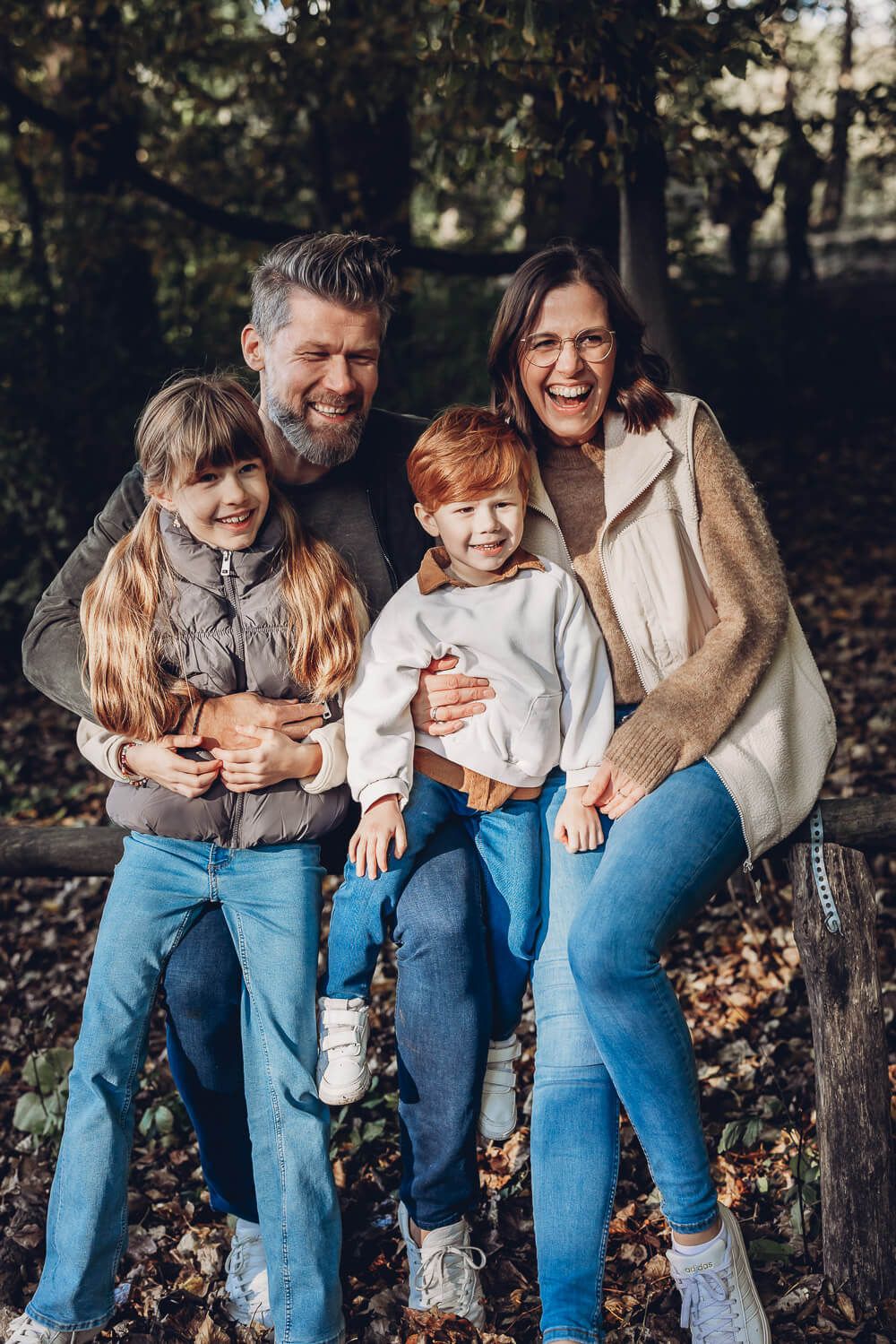 Fröhliches Familienfoto im Herbst in Hannover – Eltern sitzen mit zwei Kindern auf einer Bank im Wald und lachen gemeinsam