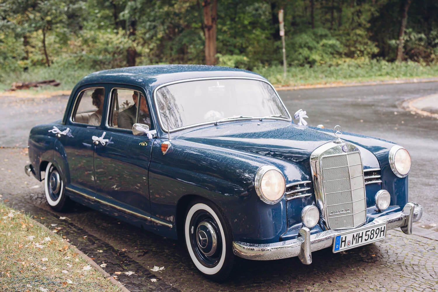 Geschmückter blauer Hochzeitsoldtimer mit Brautpaar auf regennasser Straße bei einer Hochzeit in Hannover