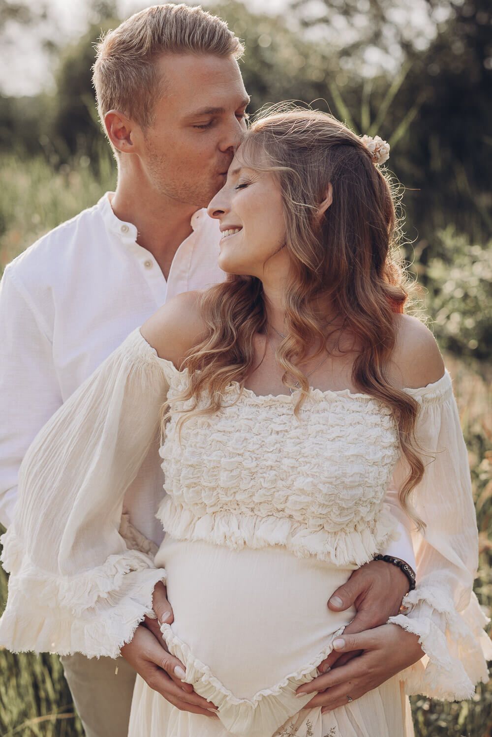 Mann küsst die Stirn der schwangeren Frau während beide sich im Grünen aneinander lehnen - Fotoshooting in Wolfsburg