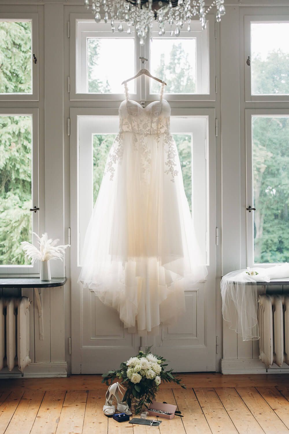 Brautkleid hängt beim Getting Ready vor großen Fenstern in einer stilvollen Hochzeitslocation - Hochzeitsfotograf Minden
