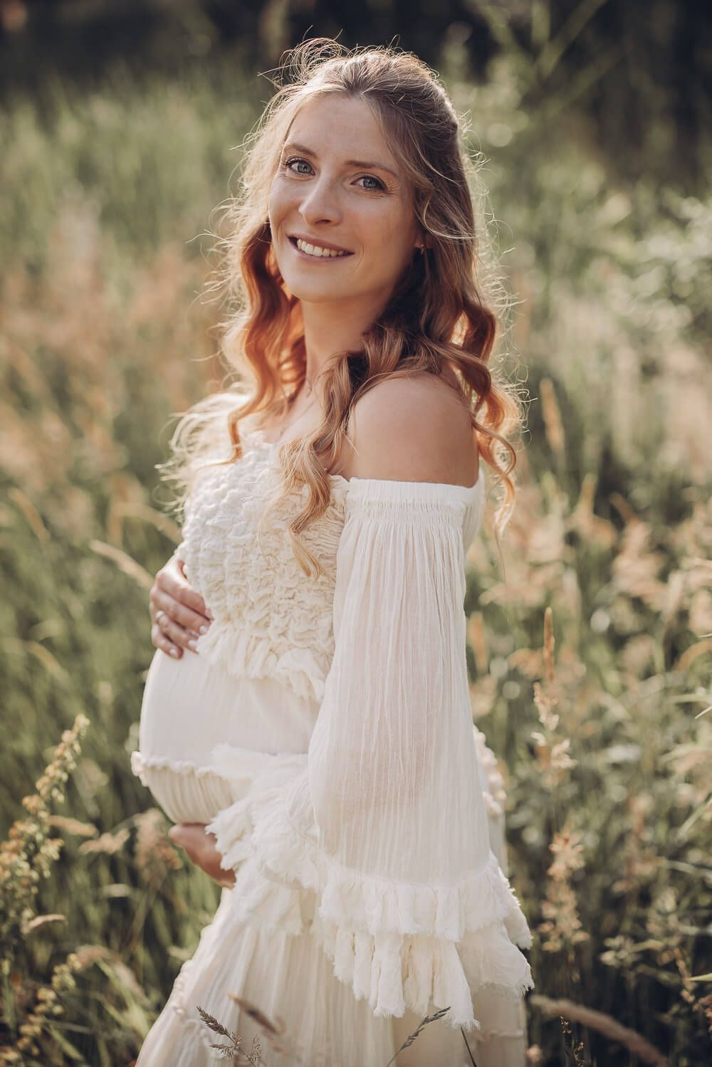 Schwangere Frau im weißen Kleid steht in einer sommerlichen Wiese und hält liebevoll ihren Babybauch - Outdoorshooting in Hannover
