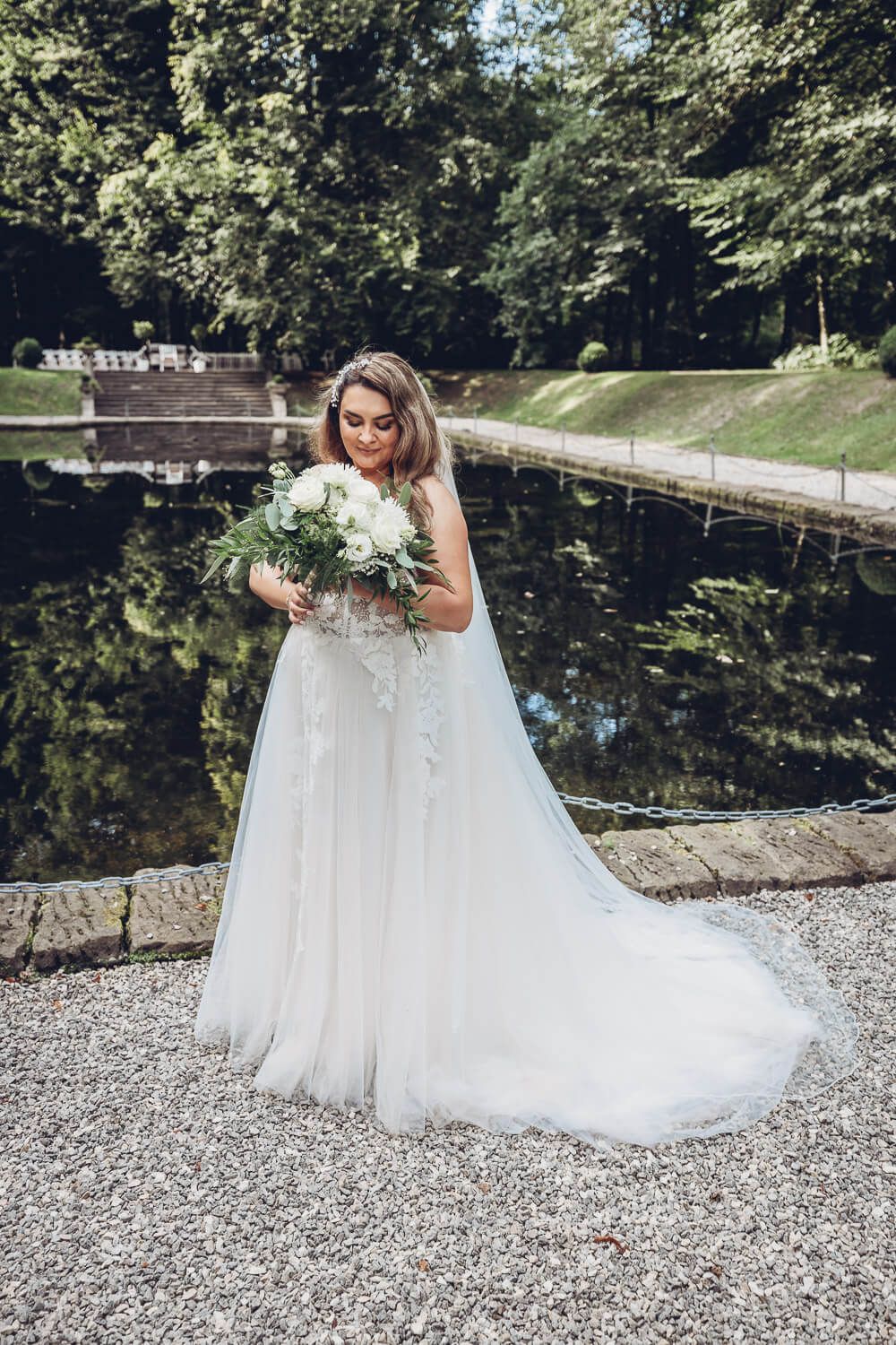 Elegantes Brautportrait mit weißem Brautstrauß am Wasser in einer grünen Parkanlage, fotografiert von einem Fotografen in Hannover