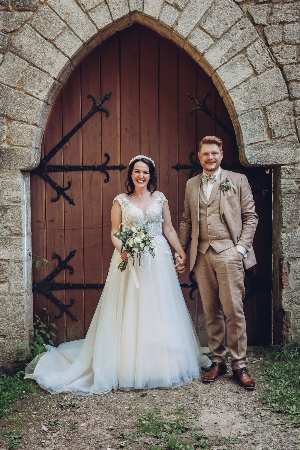 Brautpaar steht Hand in Hand vor einem historischen Holztor und lächelt bei seinem Hochzeitsportrait